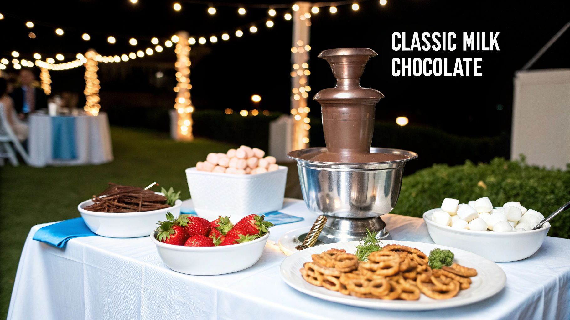 A chocolate fountain with strawberries, pretzels, and marshmallows at an outdoor evening event.