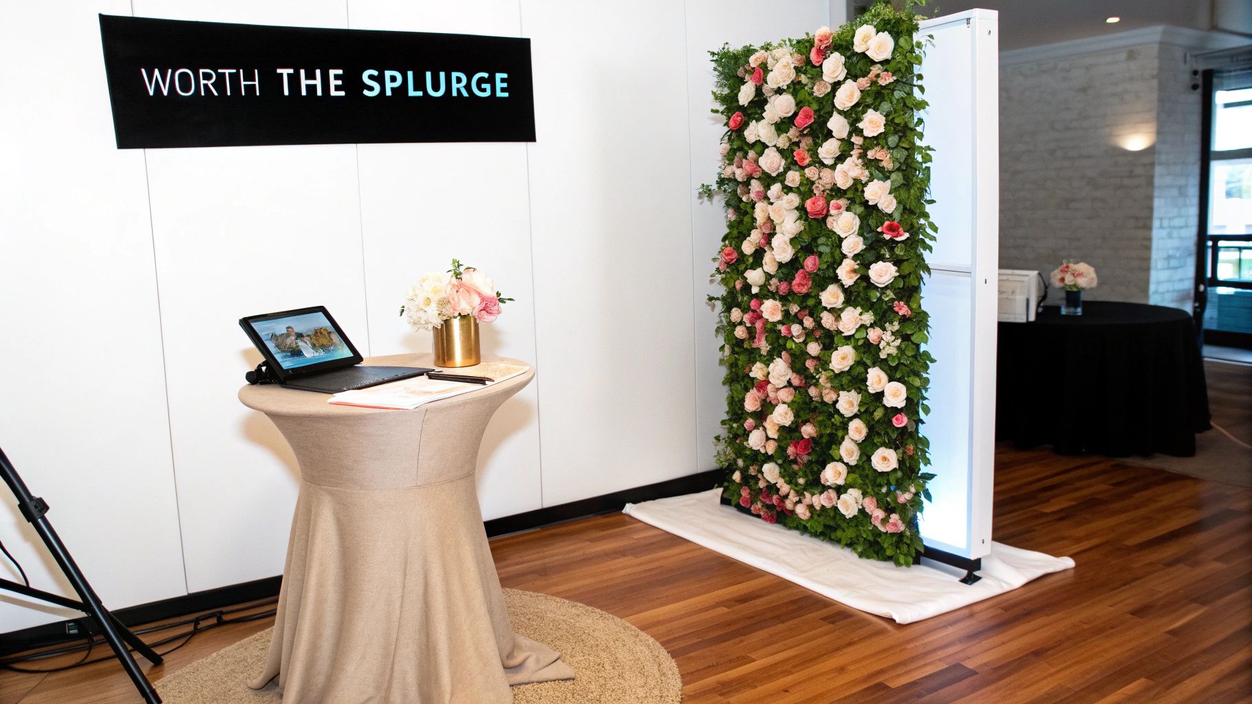 A stylish event photo booth setup featuring a flower wall and a tablet stand with a sign.