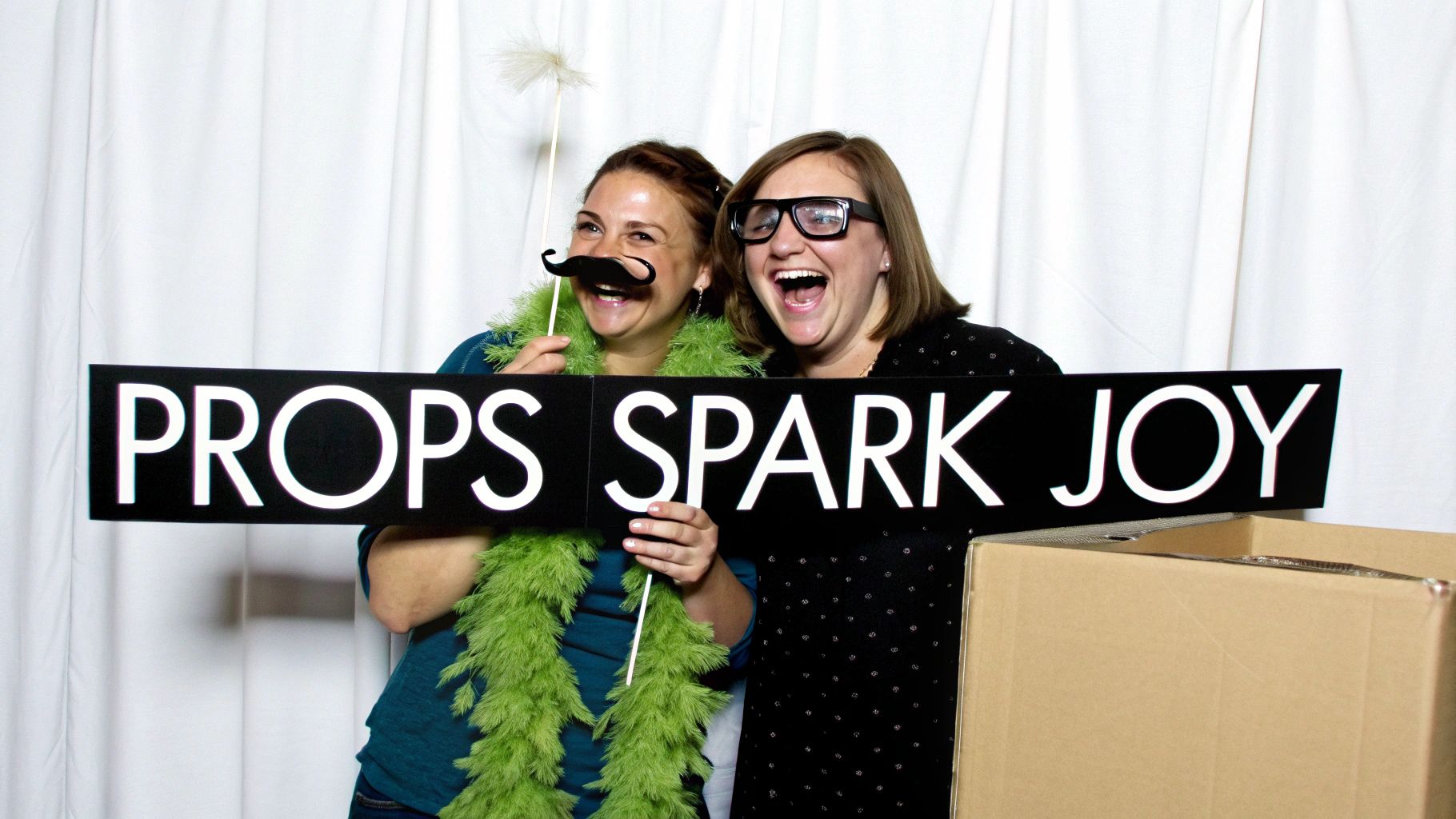 Two women laughing with photo booth props holding sign that says props spark joy