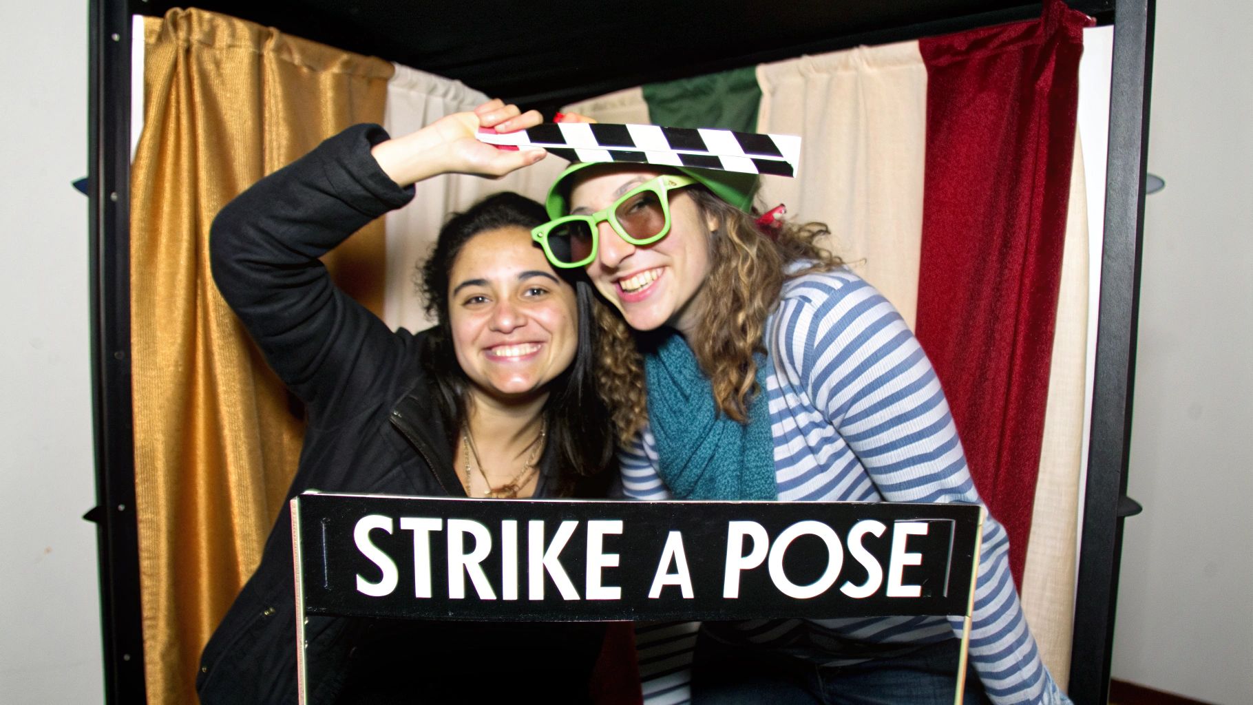 Two smiling women in a retro photo booth with props, one wearing green sunglasses and a hat.