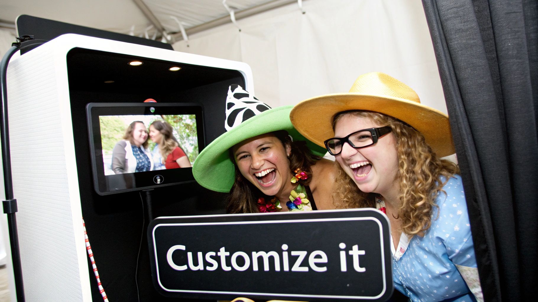 Two happy women wearing hats laughing enthusiastically while peeking out from a photo booth.