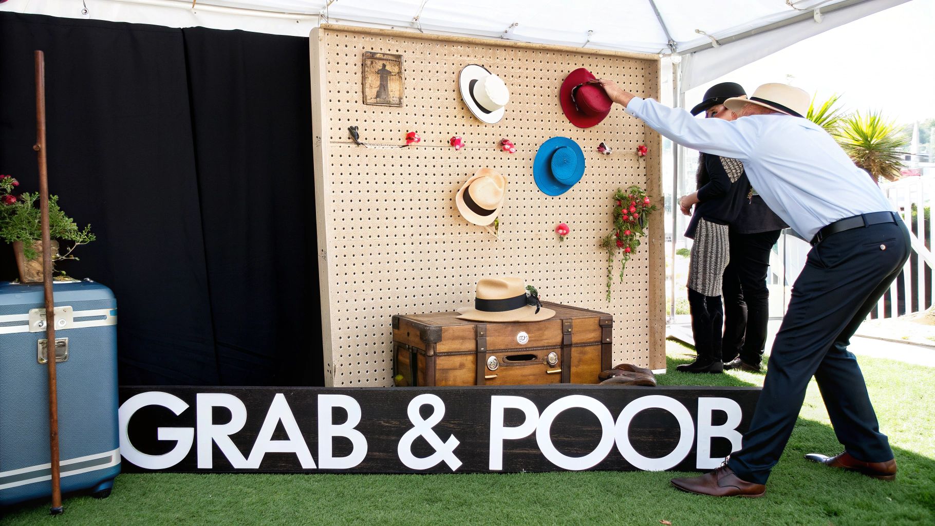 Guests select hats from a pegboard wall display at an outdoor photo booth setup on grass.
