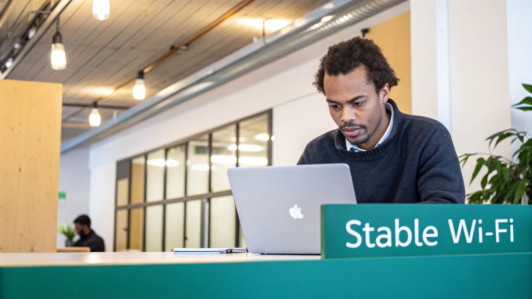 An African American man works on a MacBook laptop at a desk with a 'Stable Wi-Fi' sign in a modern office.