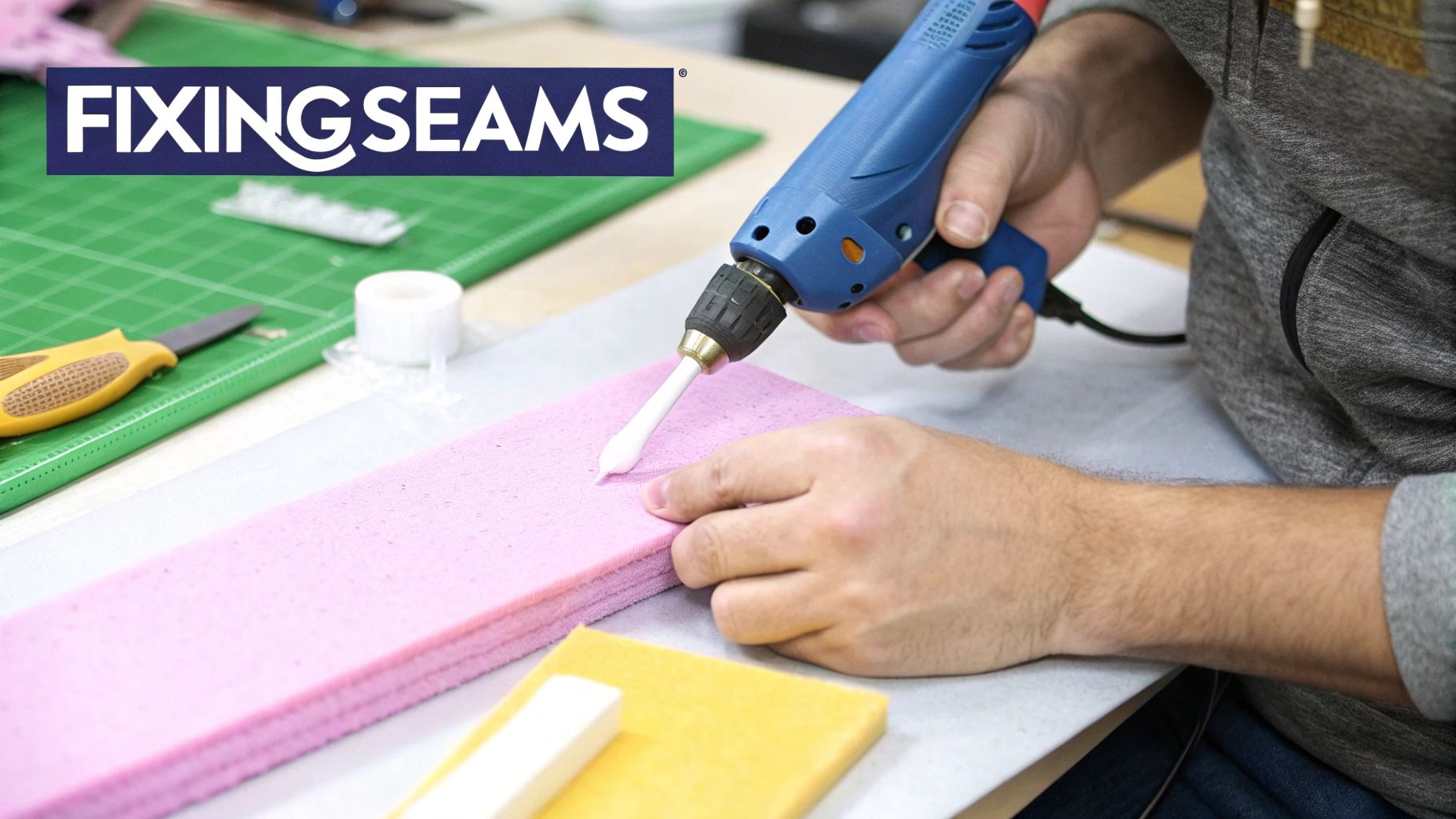 Close-up of hands using a blue tool to apply white adhesive to a pink foam sheet, amidst crafting supplies.