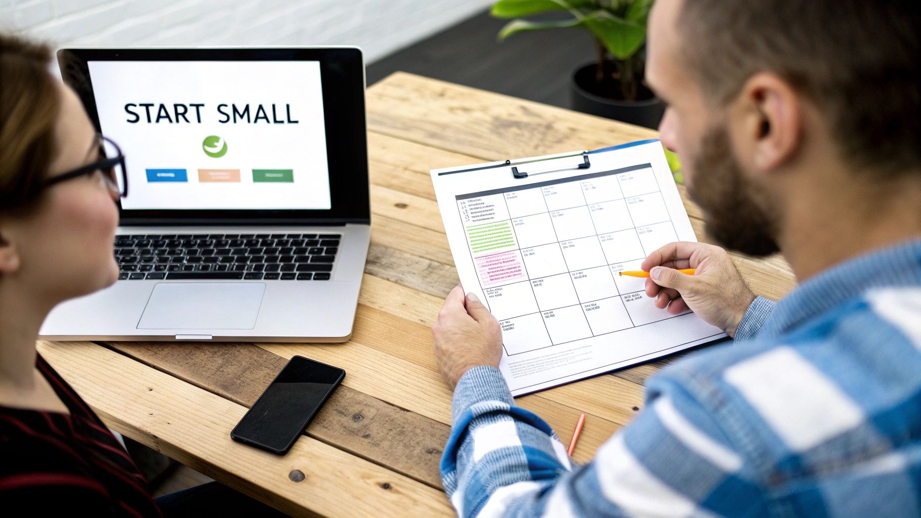 Two people collaborating at a wooden table, one looking at a 'Start Small' laptop screen, the other writing on a planner.