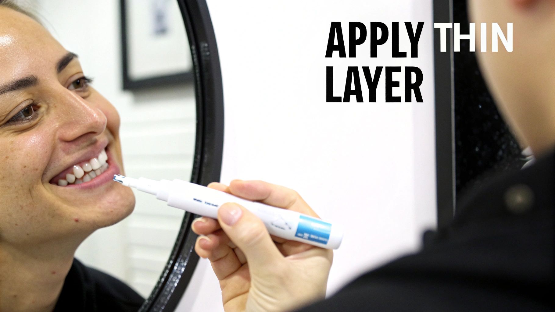 A person smiles while applying a teeth whitening gel with a pen to their lower teeth, reflected in a mirror.