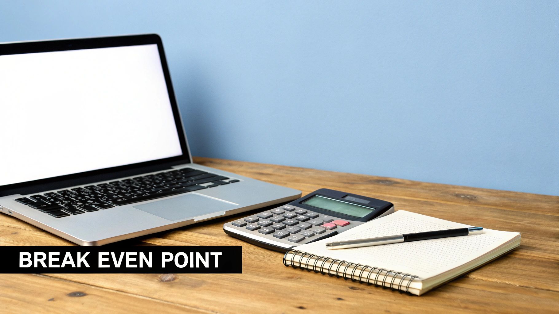 A laptop, calculator, notebook, and pen on a wooden desk, with text 'BREAK EVEN POINT'.