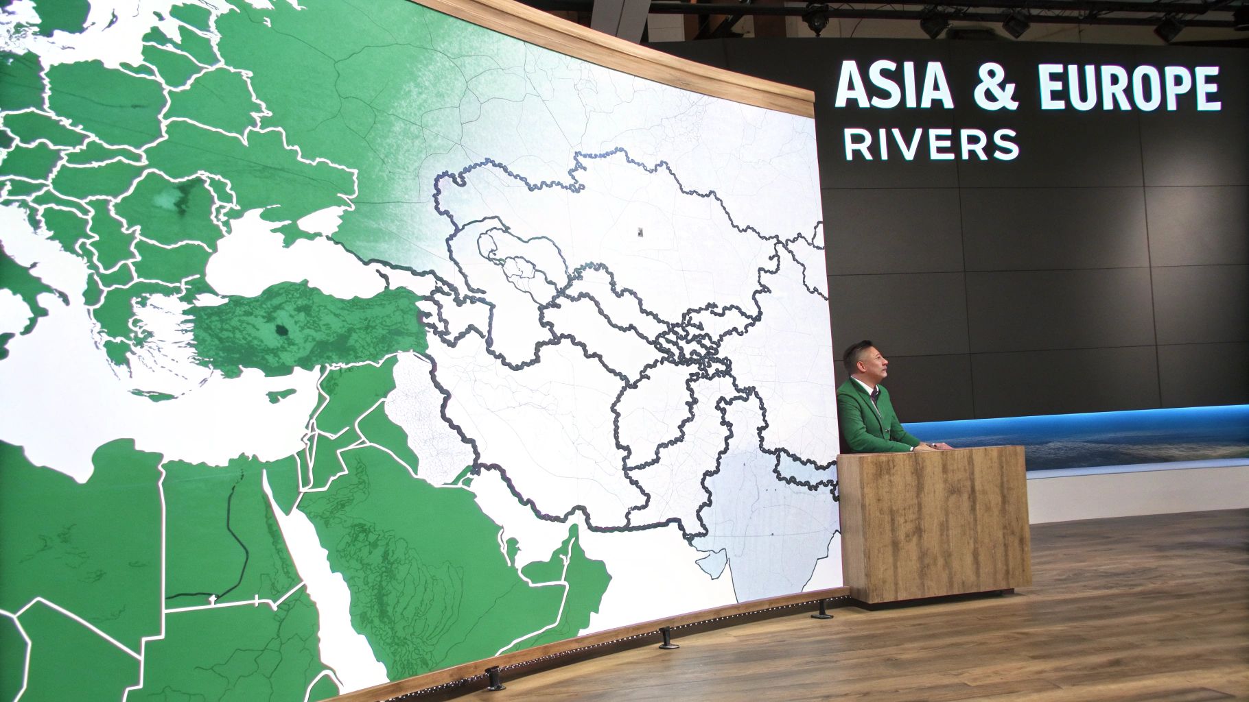A man presents a large map of Asia and Europe highlighting major rivers, standing by a podium.