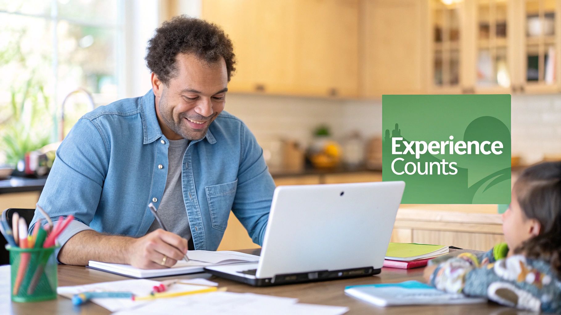 A smiling man studies at a table with a laptop, writing in a notebook next to a child.