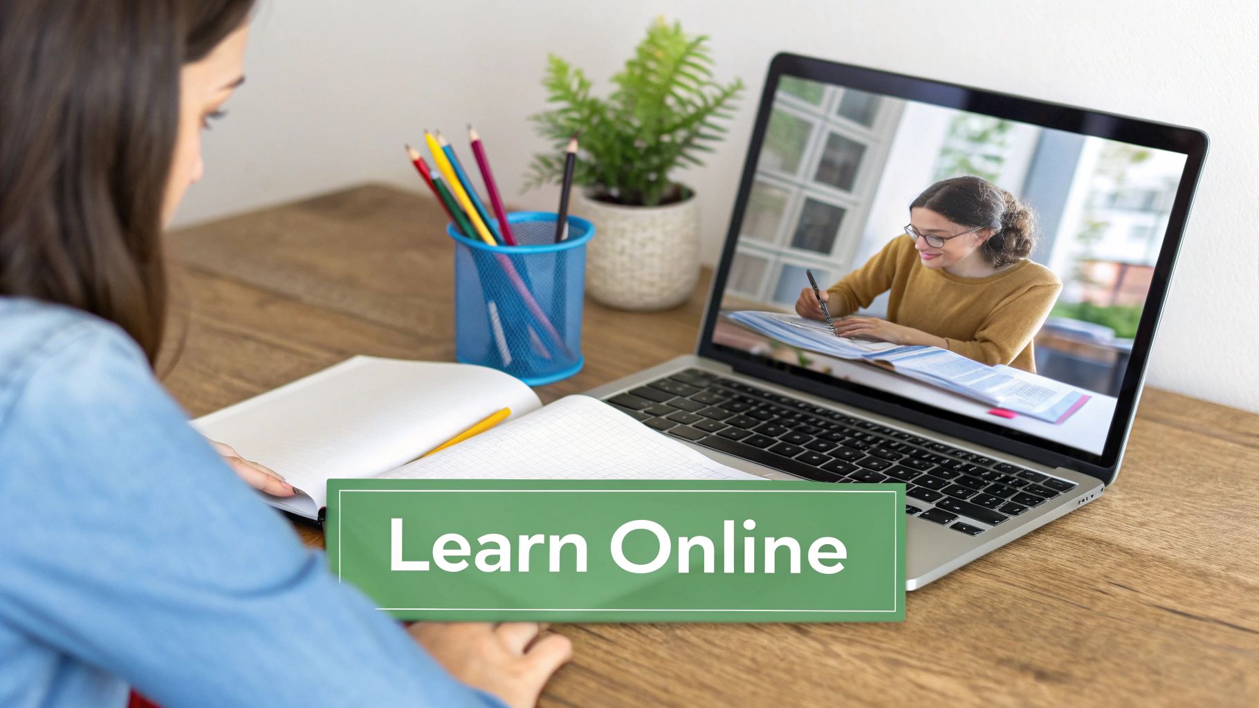 A student watches an online class on a laptop, taking notes in a notebook on a wooden desk.