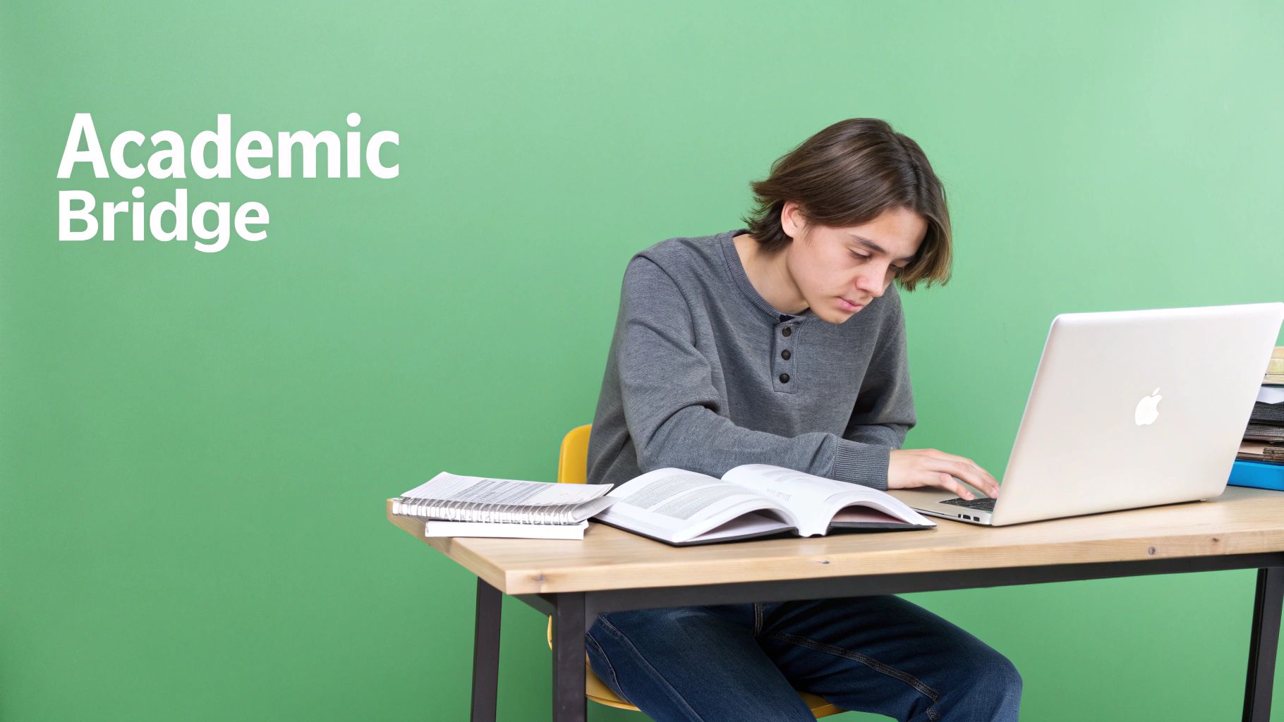 A focused student uses a laptop and studies from an open book at a wooden desk.