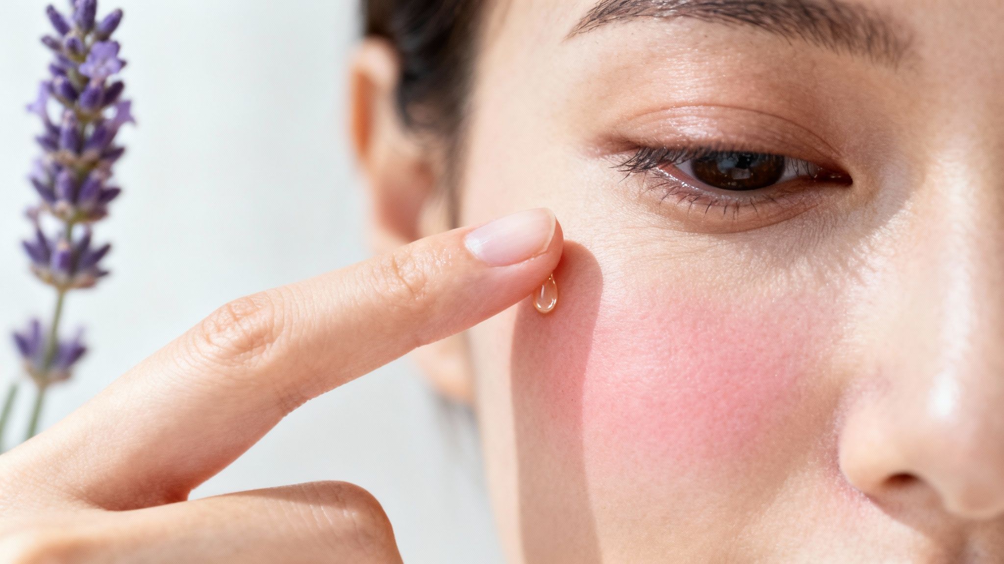 A woman applies a drop of skincare oil to her cheek, with lavender in the background.