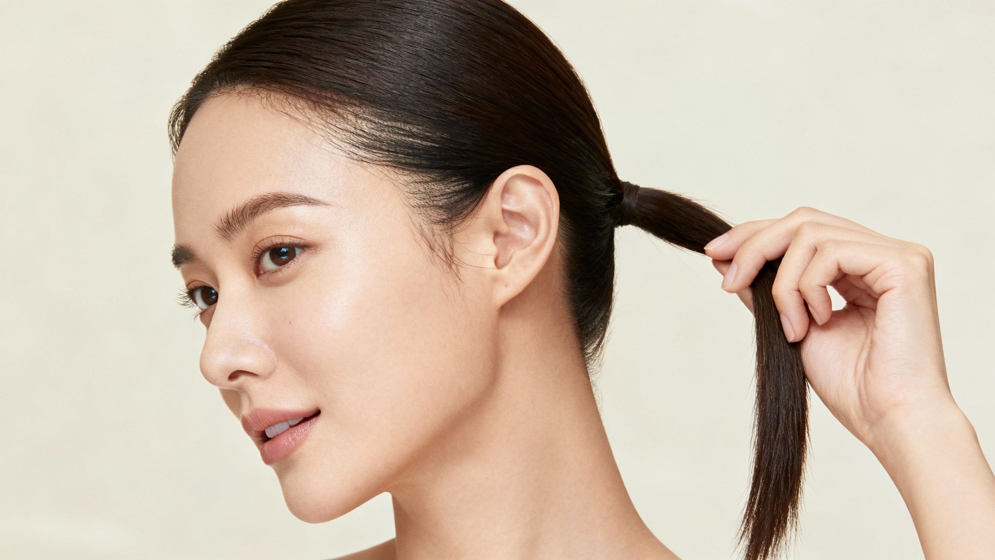 A young woman with dark hair in a low ponytail gently holds a section of her hair.