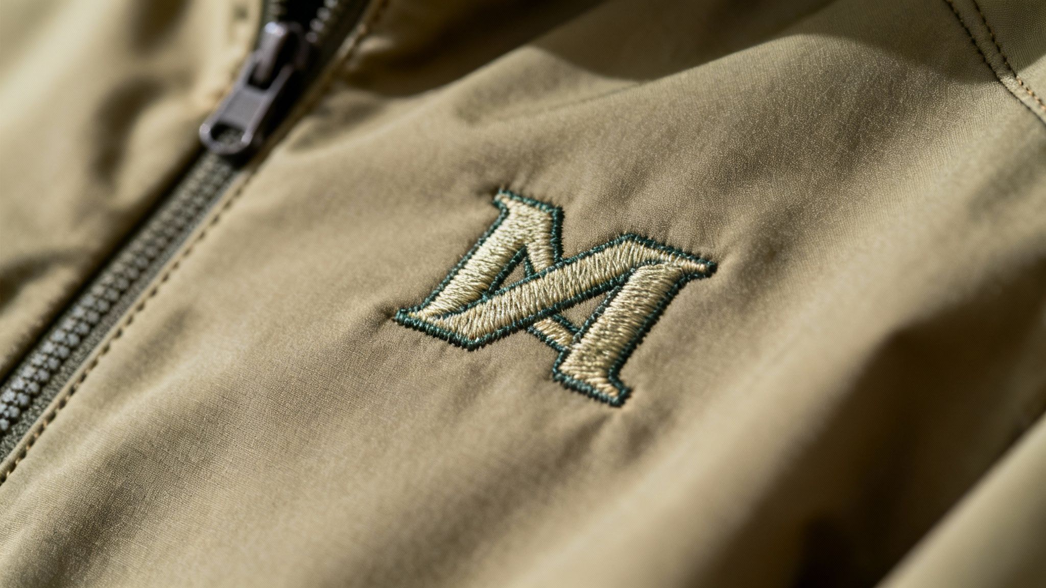Close-up of a khaki soft shell jacket featuring an embroidered MA logo and a metal zipper.