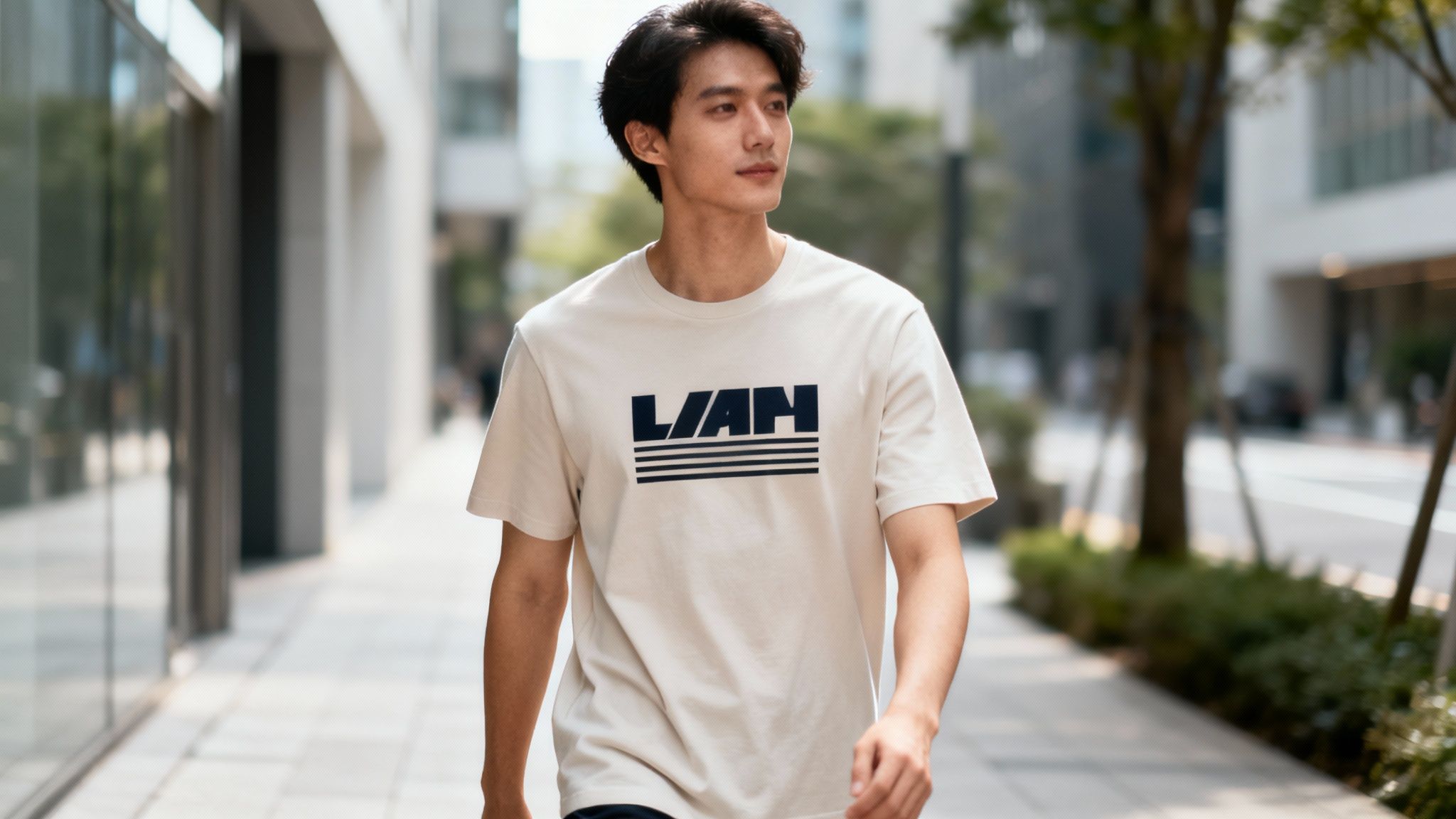A young East Asian man walks outdoors, wearing a light t-shirt with a dark 'L/AH' logo and stripes.