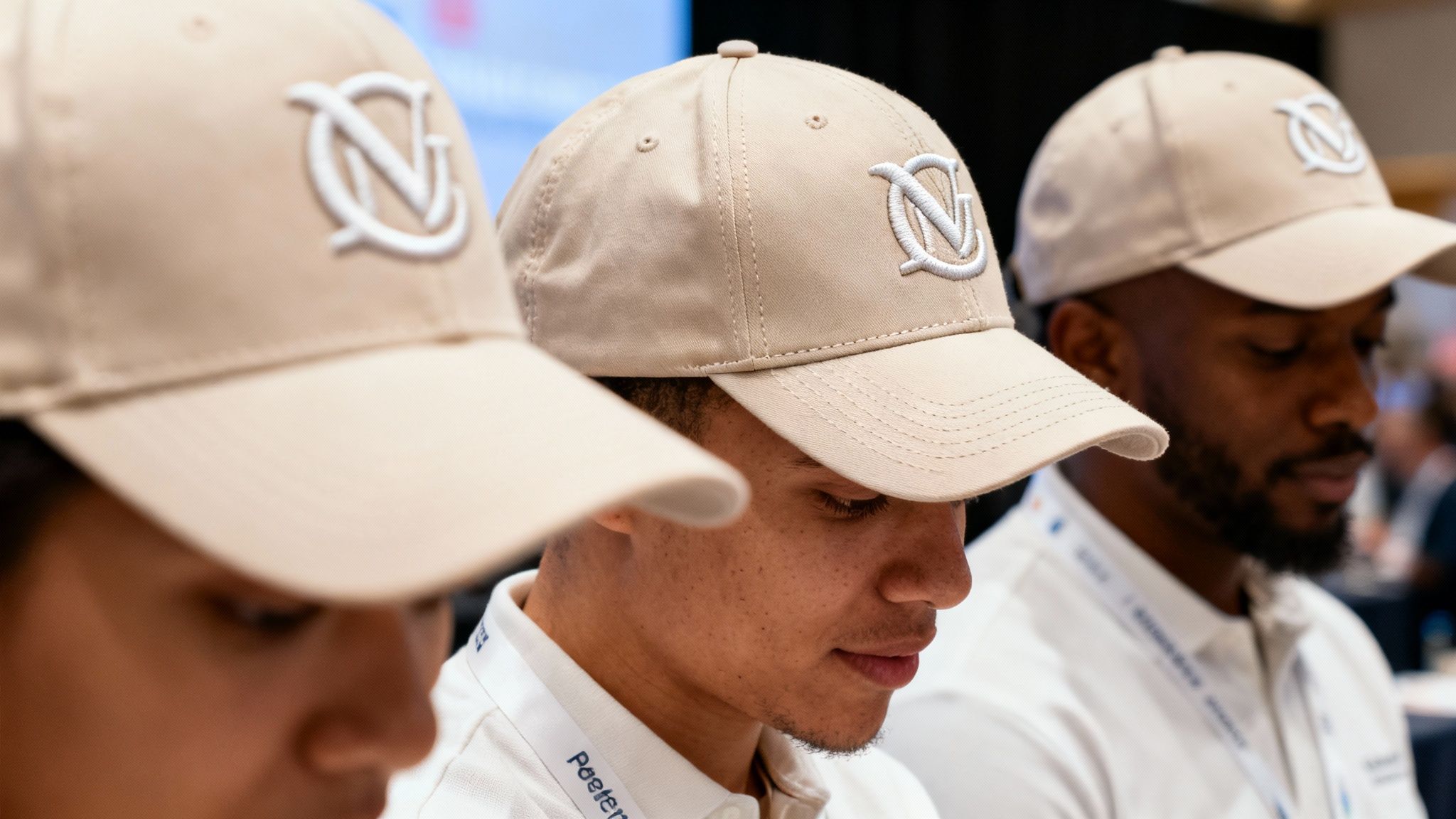Three individuals wear matching beige baseball caps with a white embroidered 'VD' or 'DN' logo at an event.