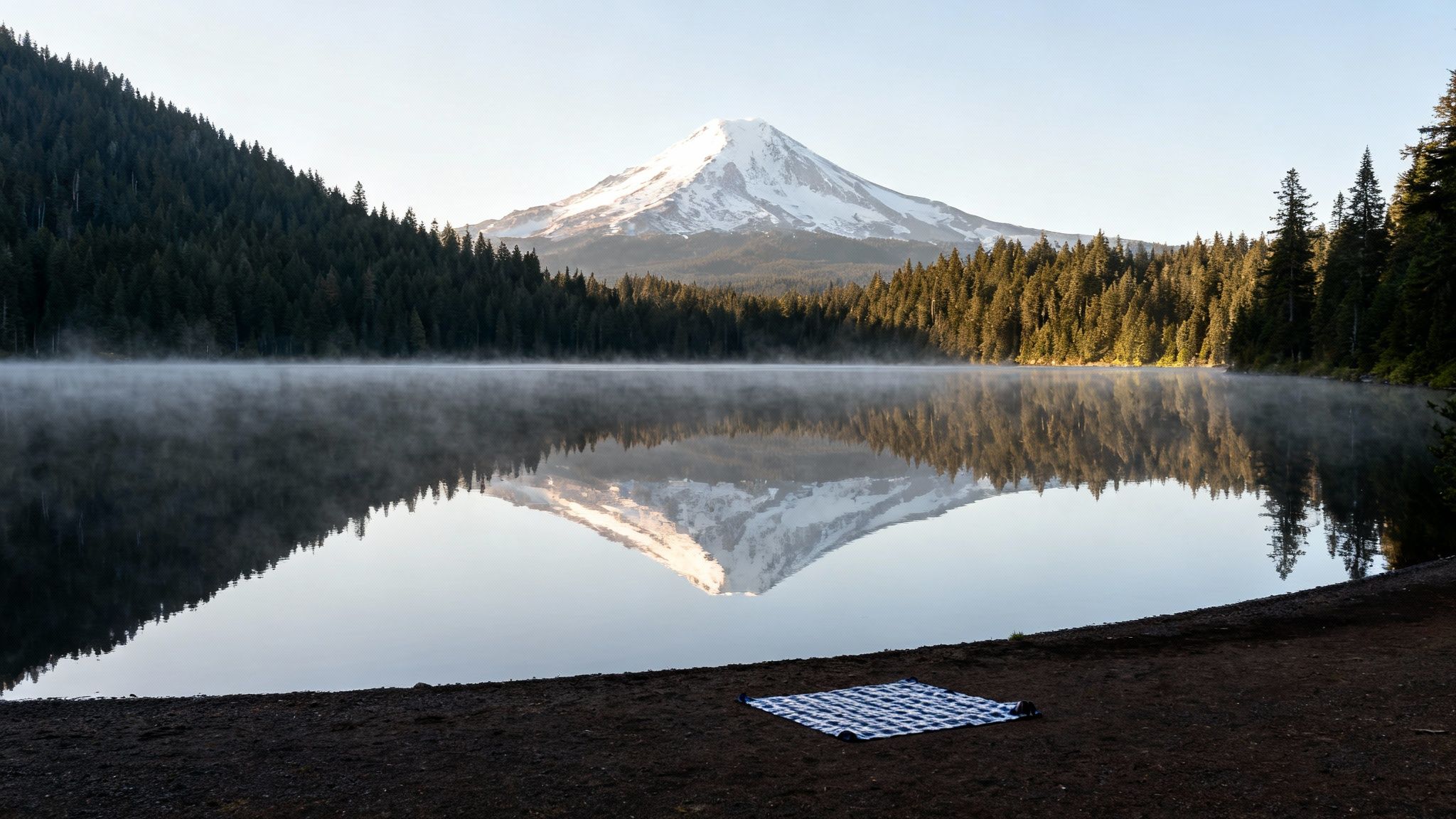 A tranquil lake reflects a snow-capped mountain and forest, with morning mist and a picnic blanket.