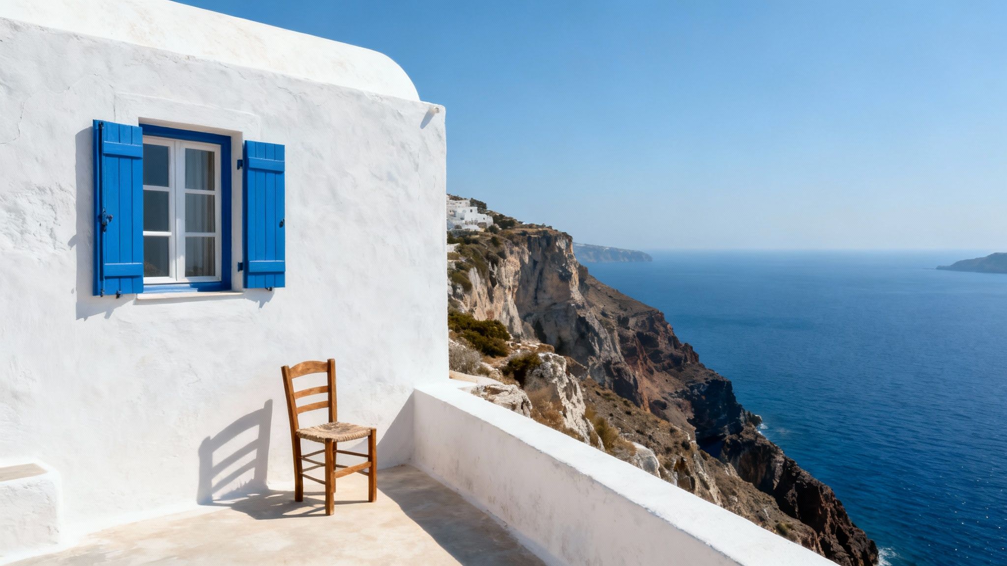 A picturesque white building with blue window shutters and a wooden chair overlooking the Aegean Sea.