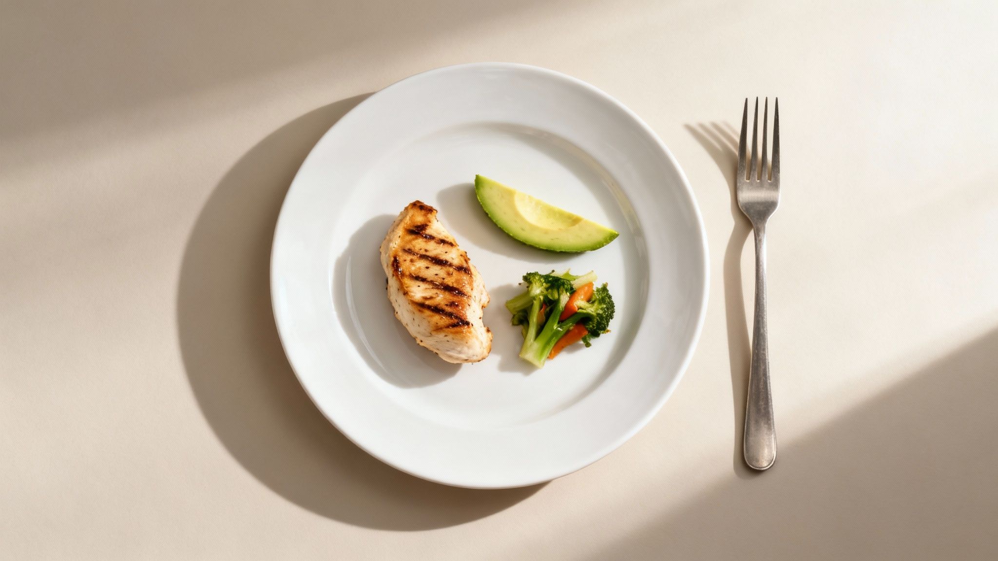 A white plate with grilled chicken breast, avocado slice, and steamed broccoli and carrots, next to a fork.