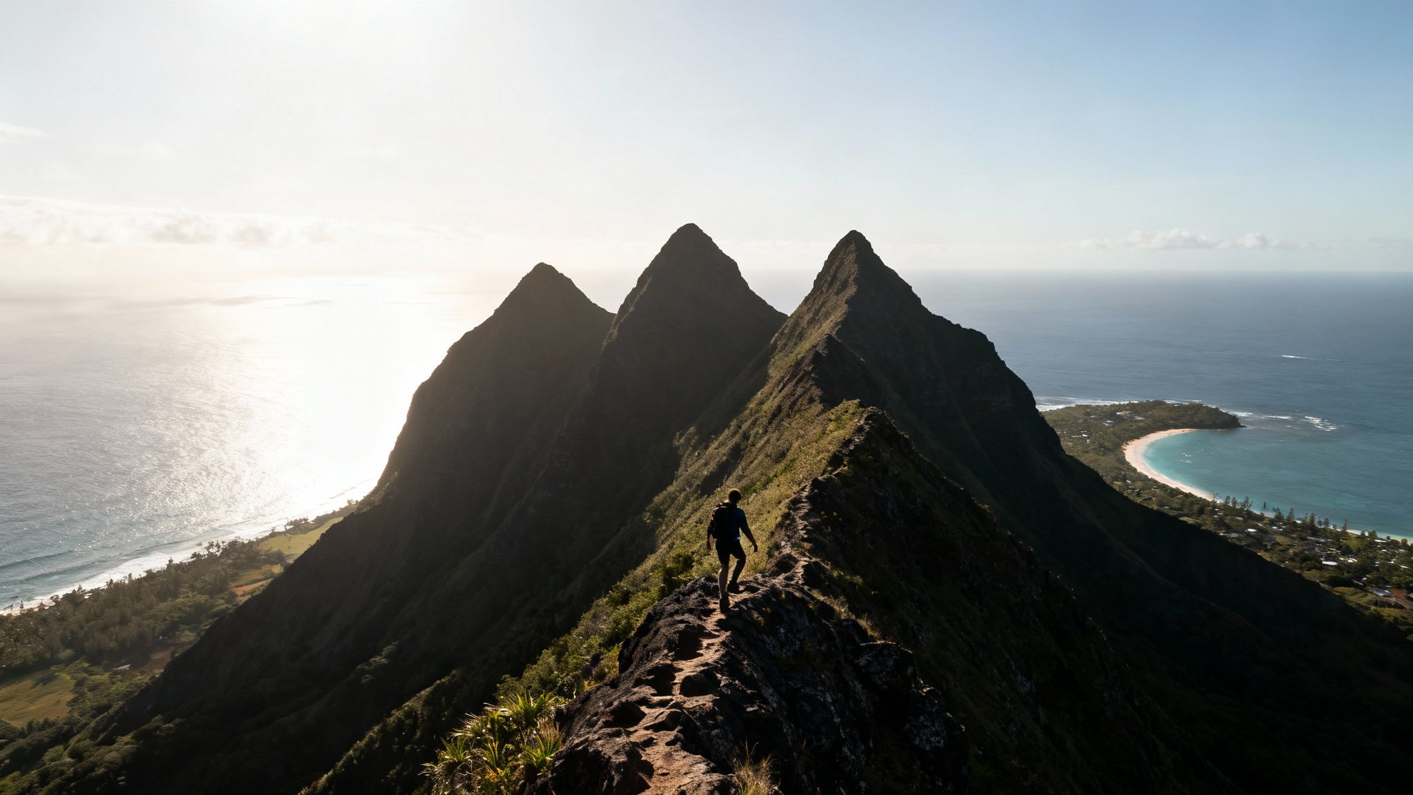 A person hikes a narrow mountain ridge with three peaks overlooking a bright ocean and a coastal town.