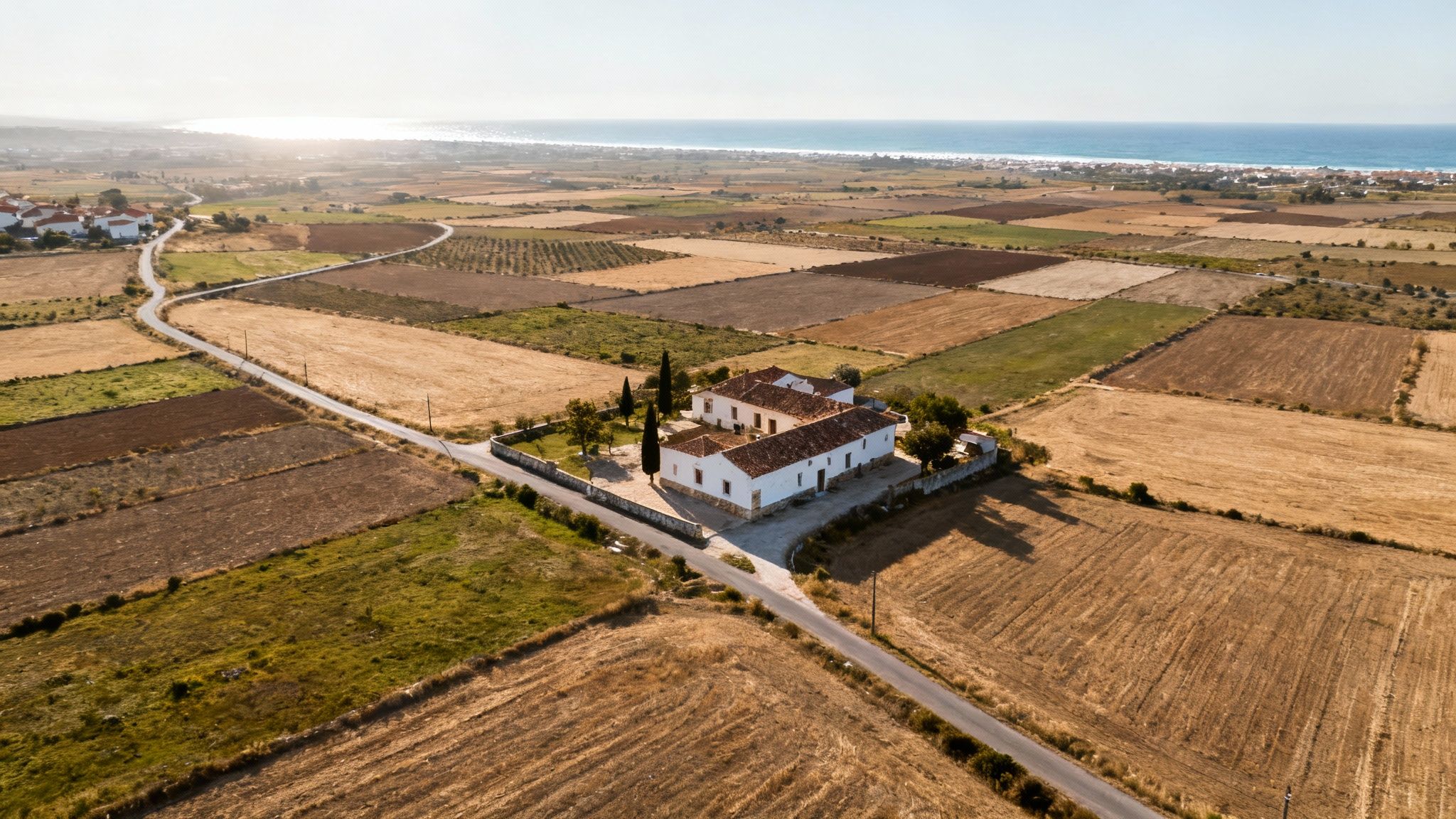Aerial view of a white traditional Portuguese farmhouse amidst golden fields, with the sea and a coastal village.