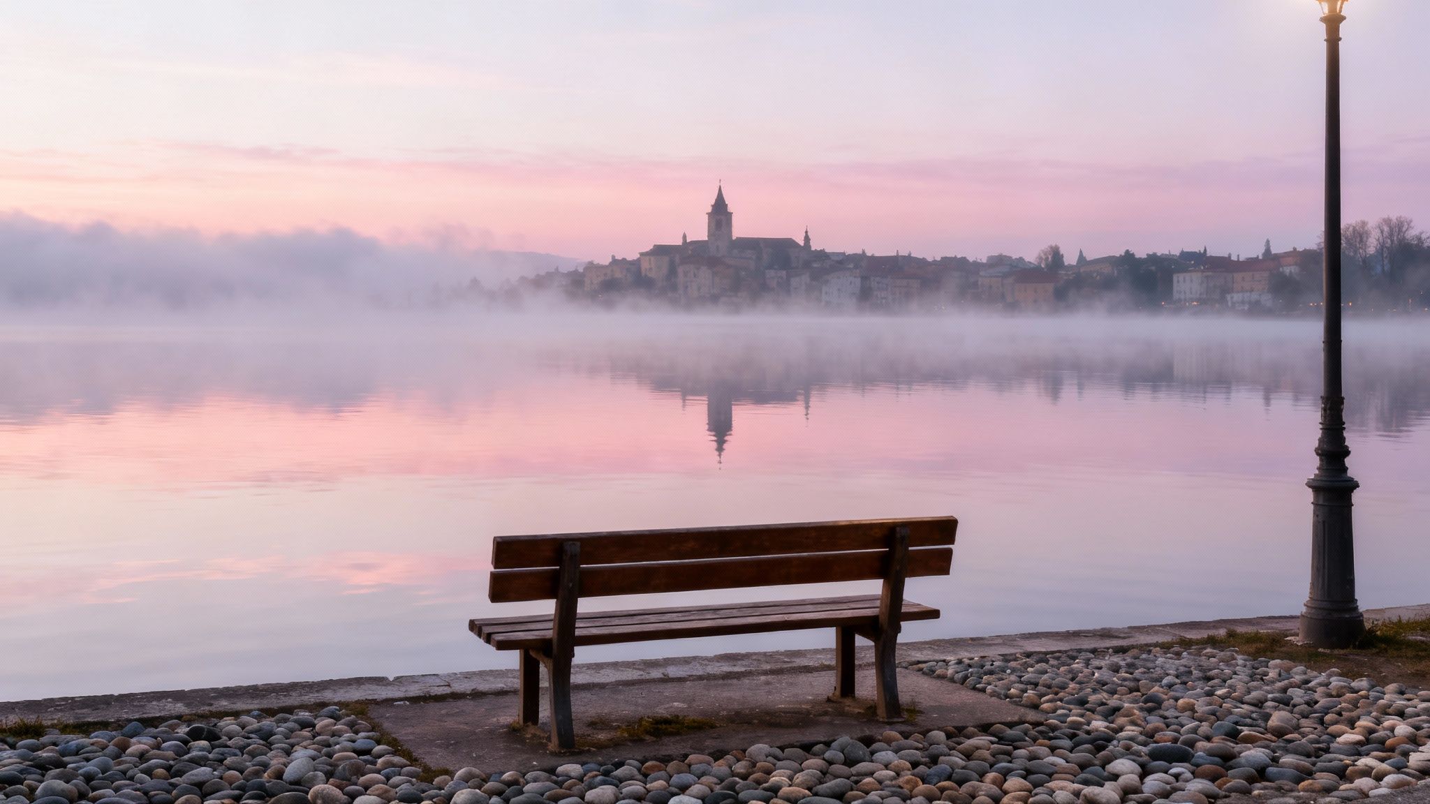 A serene morning mist over a lake with a historic town reflecting in the pink water, featuring an empty wooden bench.
