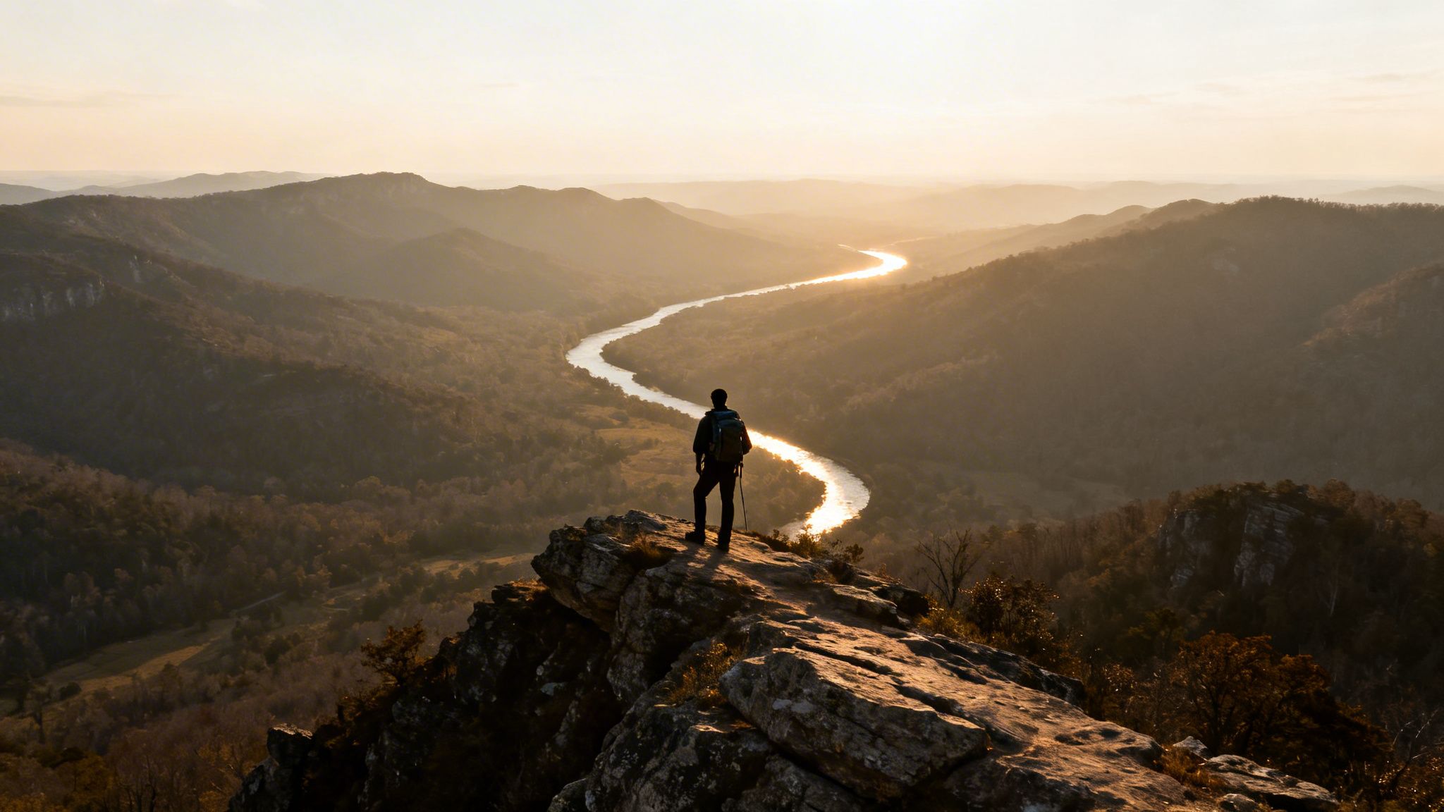 A lone hiker stands on a rocky cliff, overlooking a winding river reflecting the golden sunset amidst a vast mountain landscape.
