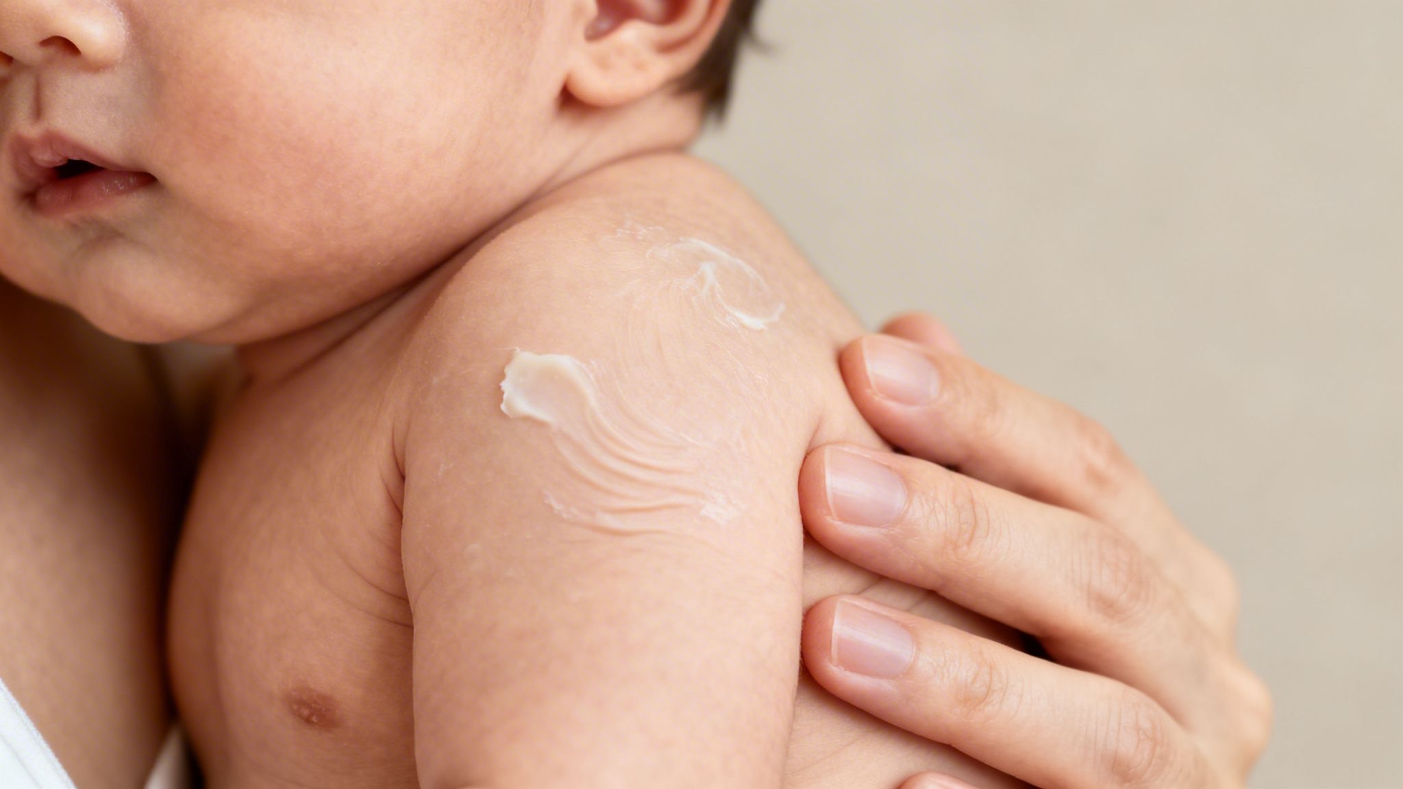 Close-up of a baby's shoulder with white cream applied, held by an adult hand.