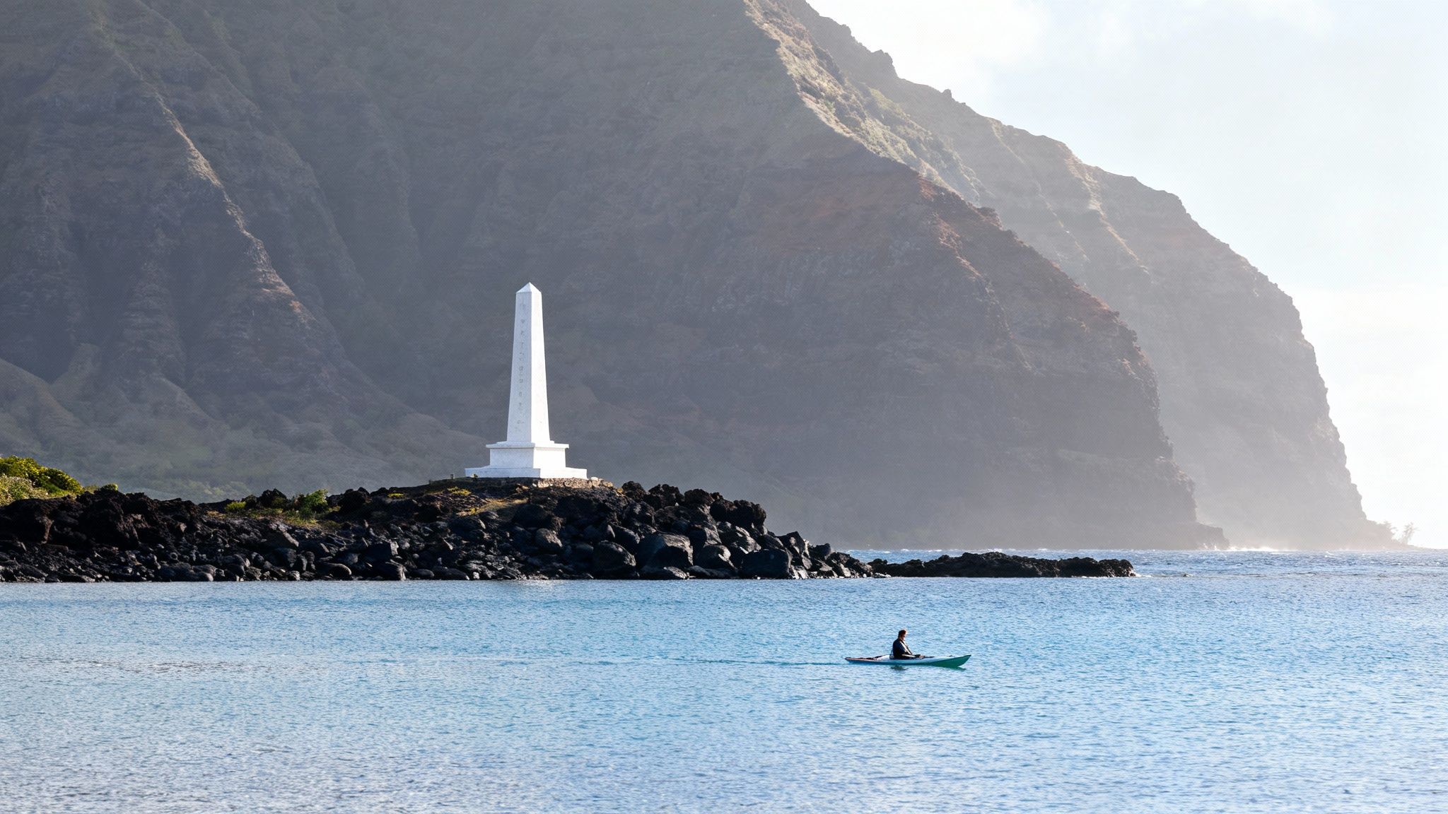 A person kayaks in blue water near a white monument on a rocky shore with misty mountains.