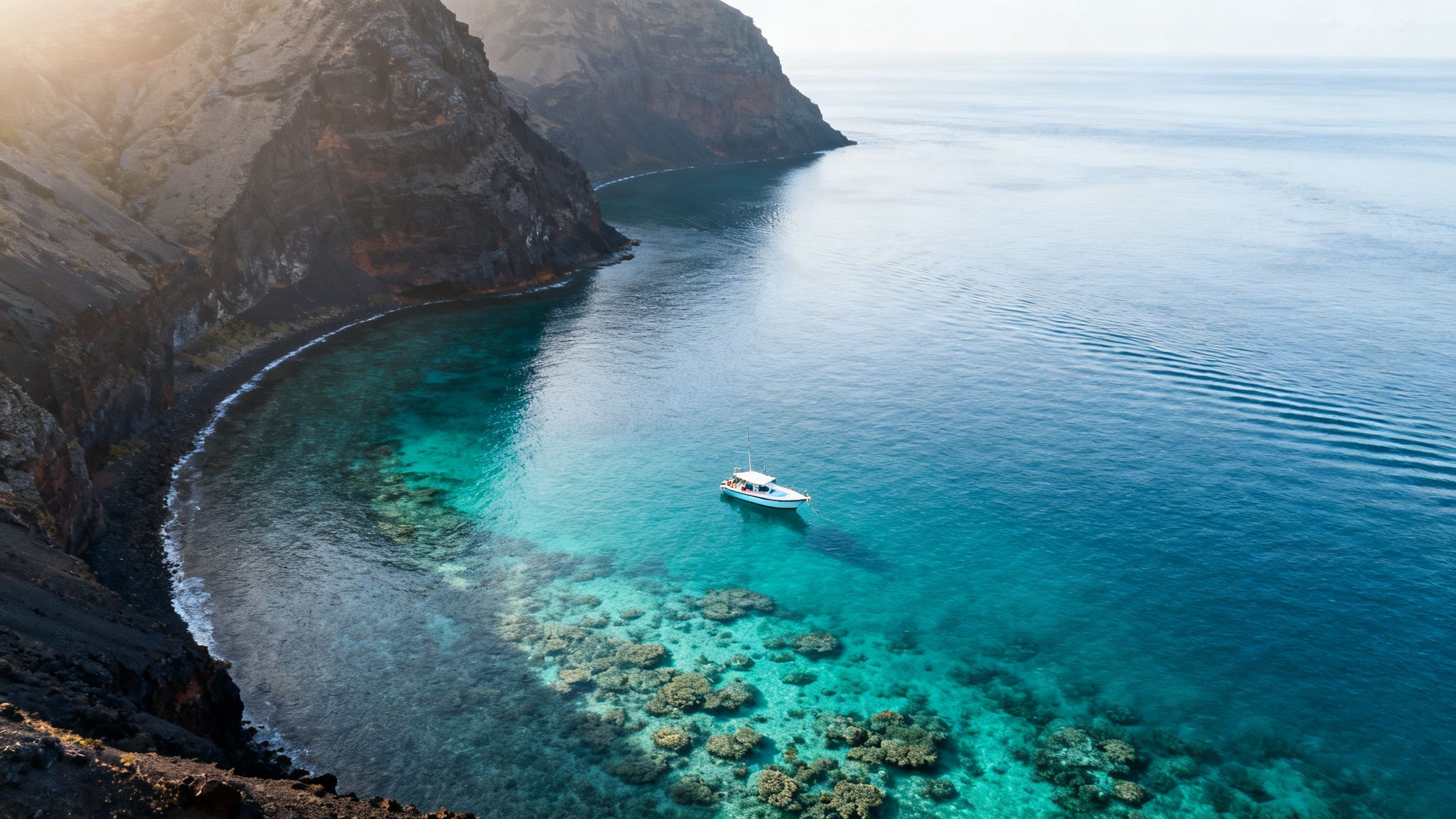 Aerial view of a white boat anchored in a clear turquoise bay surrounded by dark volcanic cliffs.
