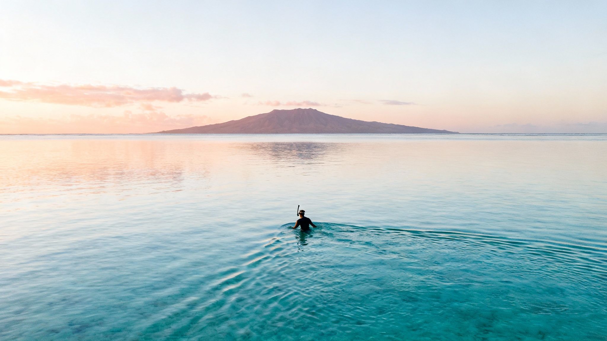 Person snorkeling in calm turquoise water with a distant island under a serene sunset sky.