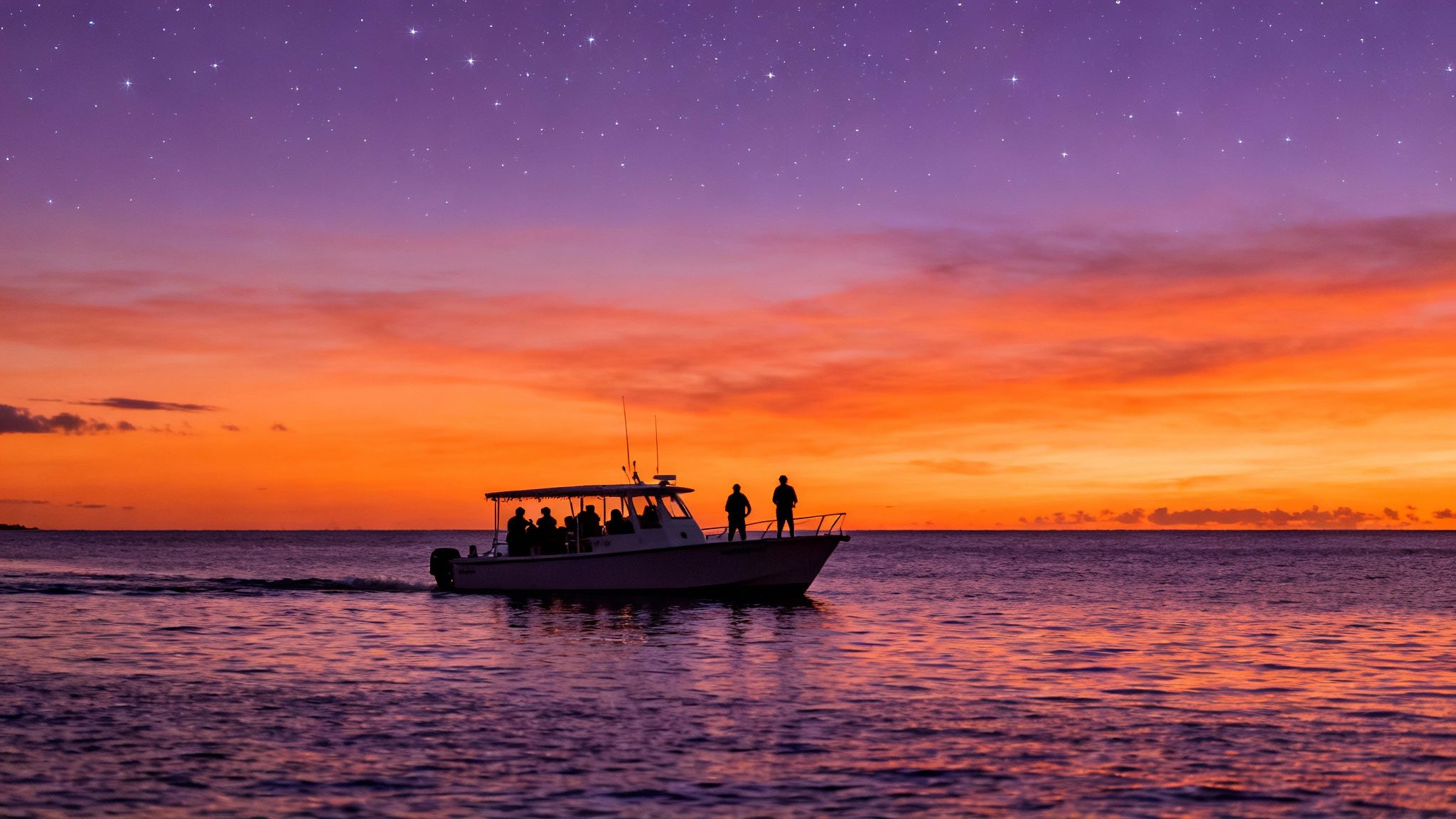 Silhouetted boat with people on calm ocean water under a vibrant sunset sky with stars.