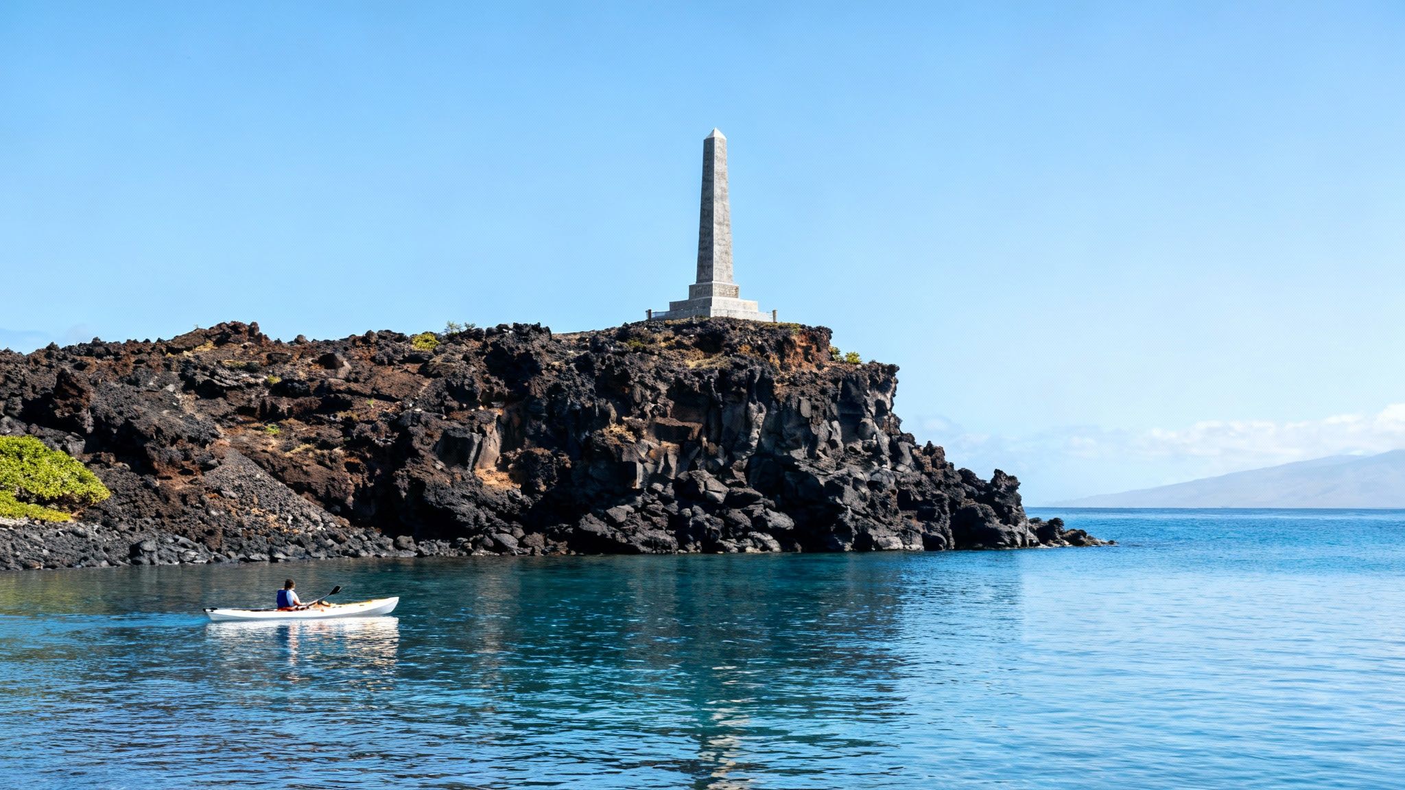 A white monument stands on the rocky shore of a clear blue bay, with lush green cliffs in the background