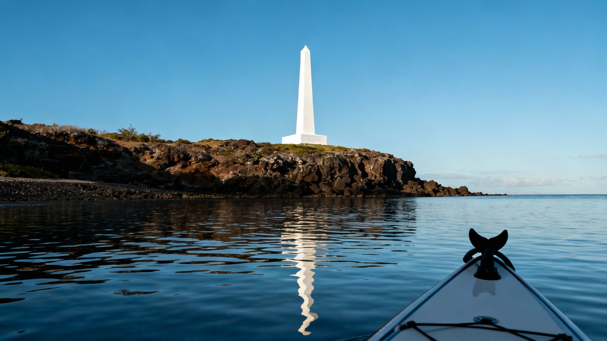 A white obelisk monument stands on a rocky island, reflecting in calm ocean water from a kayak.