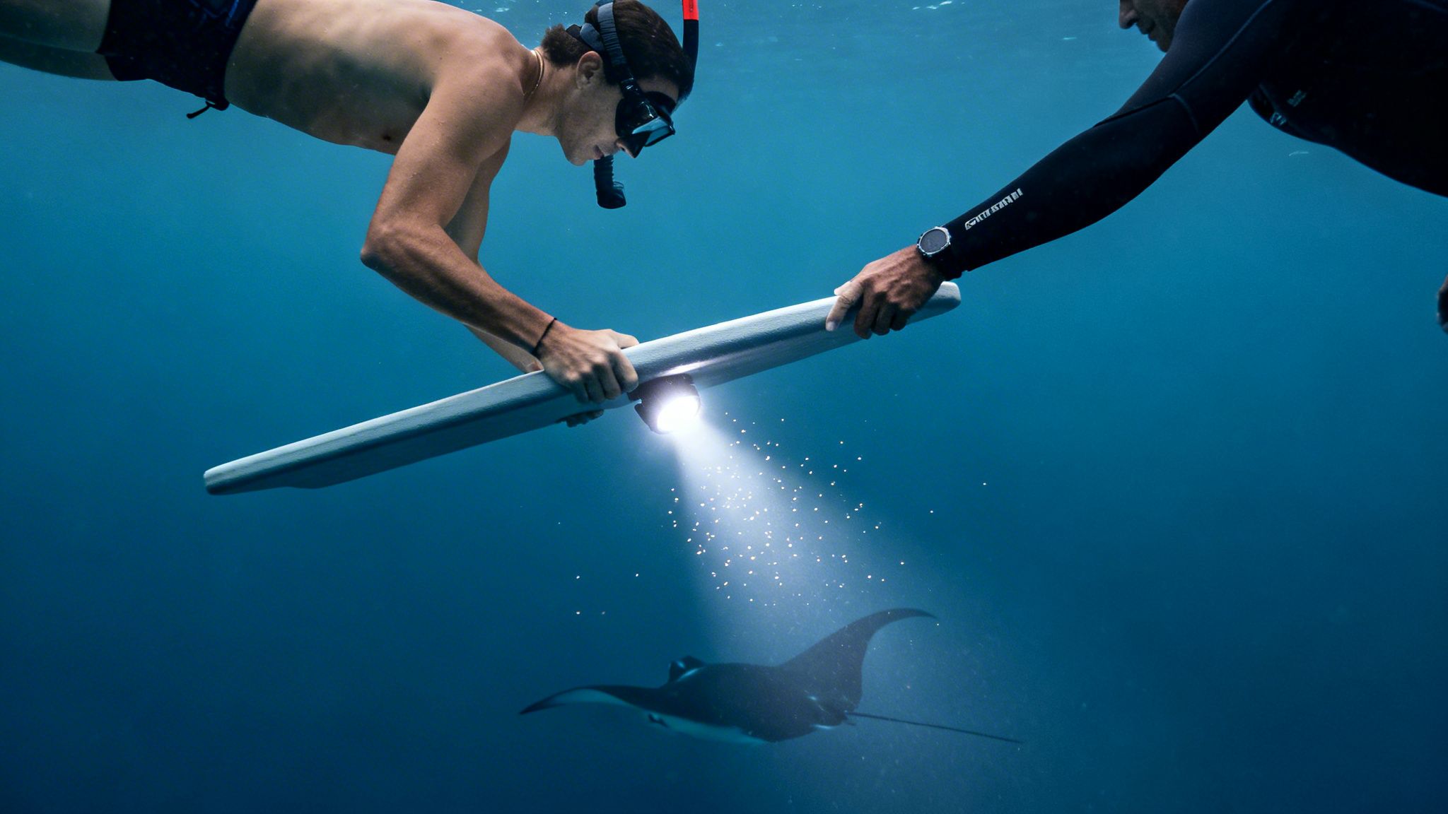 Two snorkelers illuminate a majestic manta ray with an underwater light board in deep blue waters.