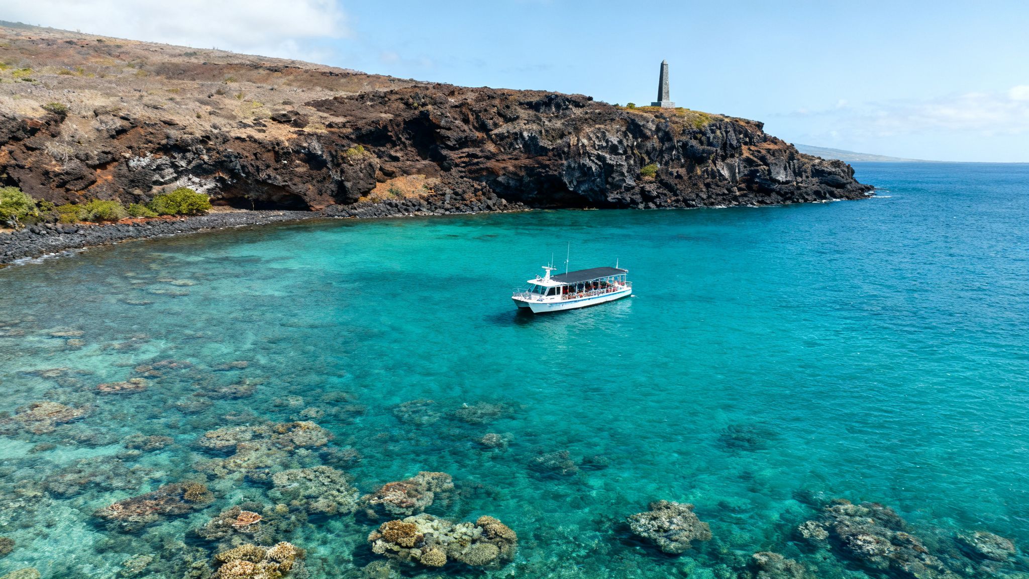 An aerial view of a tour boat in a clear turquoise bay with a coral reef and rocky coast.