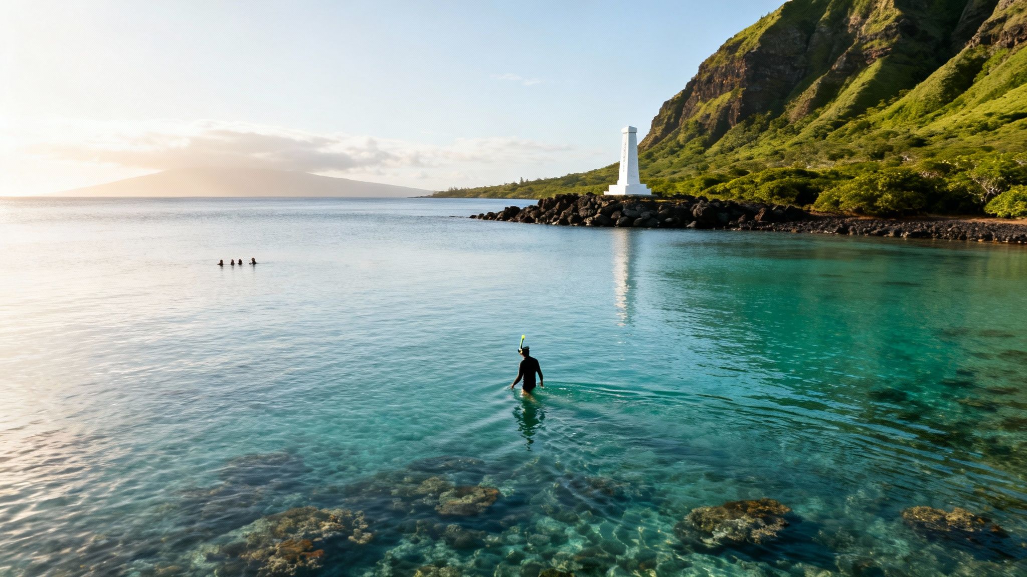 A person in snorkeling gear stands in clear tropical water near a lighthouse and lush green mountains.