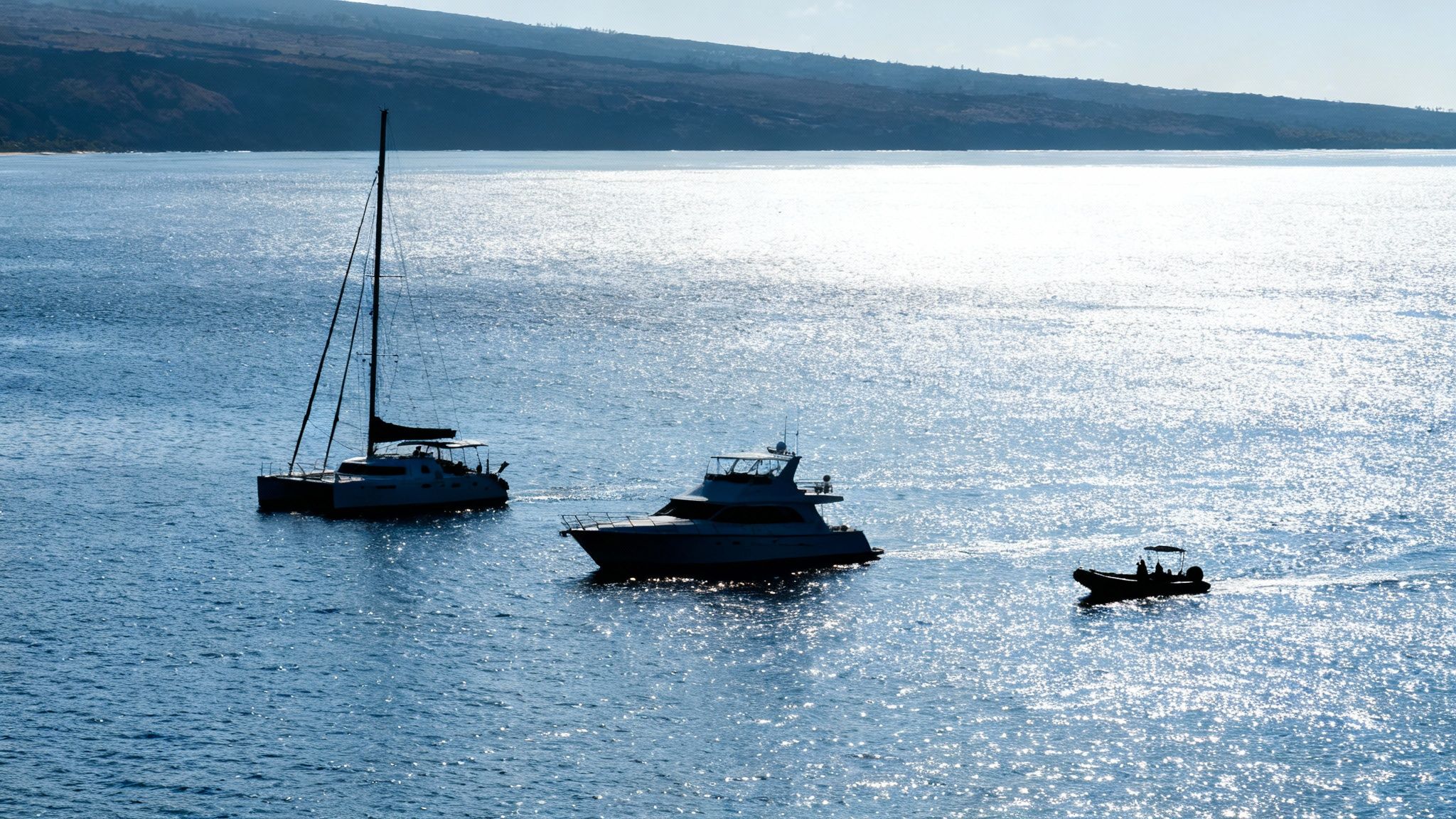 Three boats, a catamaran, yacht, and small boat, silhouetted on sparkling blue ocean with a distant coastline.