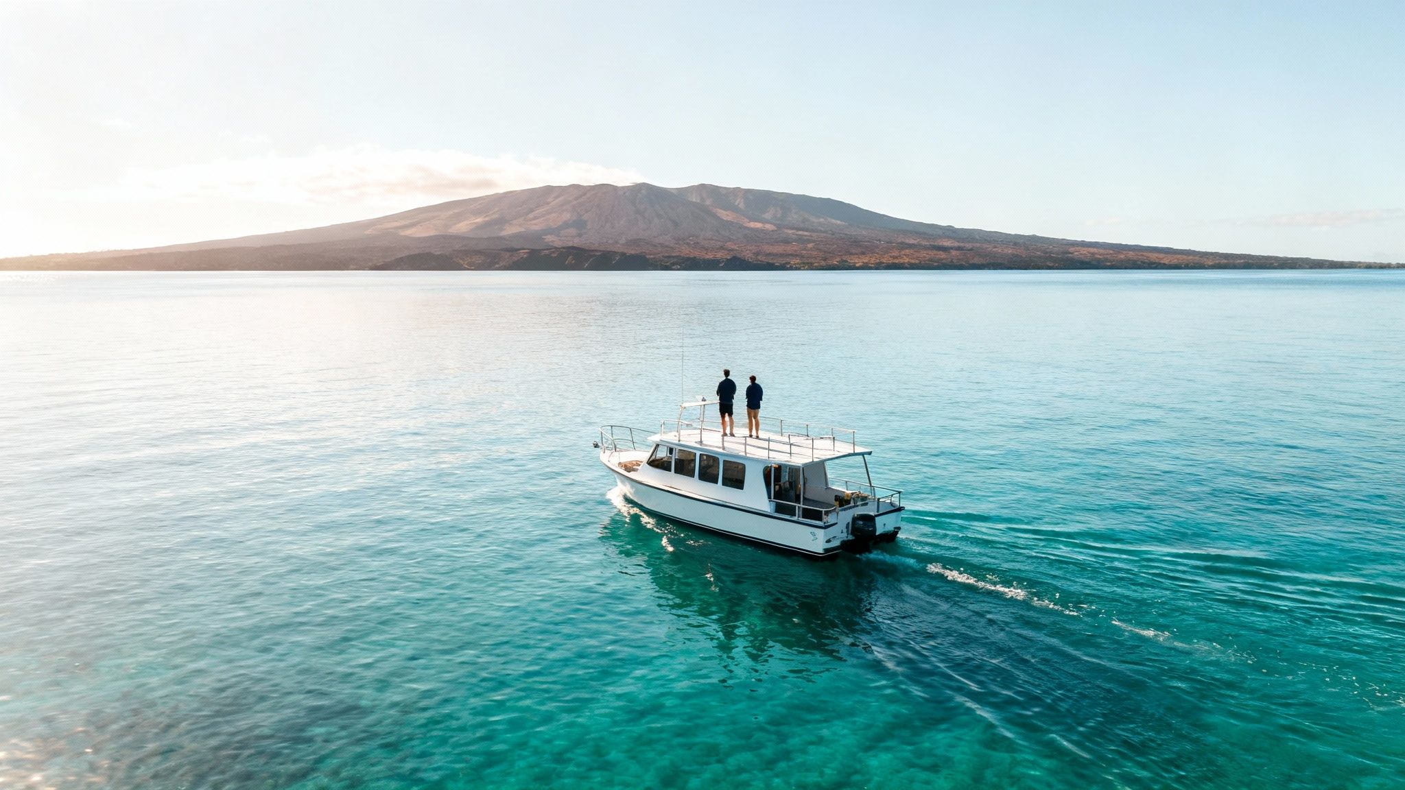 Two people stand on a boat cruising through turquoise water towards a distant island mountain.