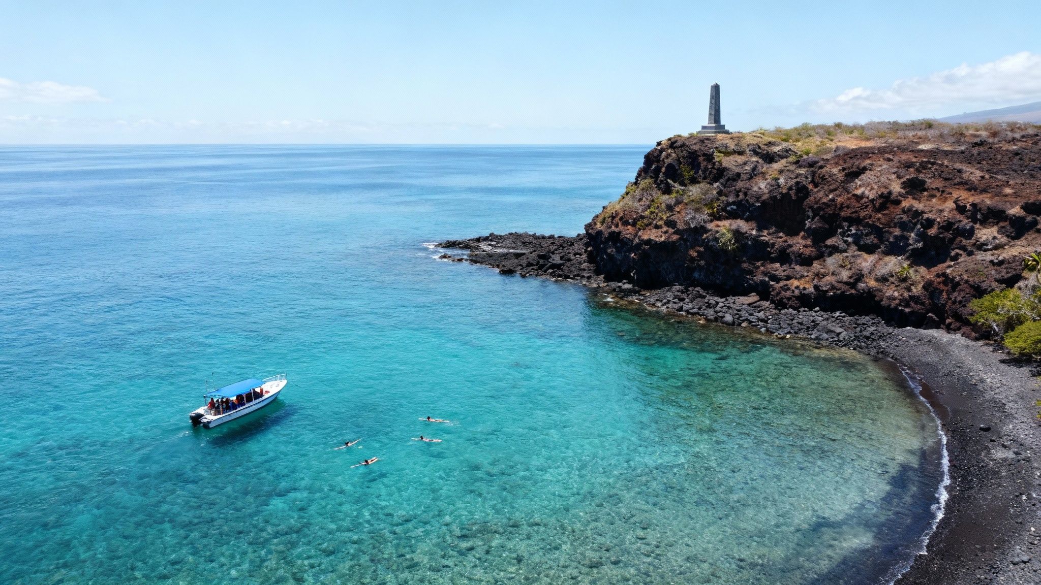 Aerial view of a boat and snorkelers in the turquoise waters near a rocky coast with a monument.