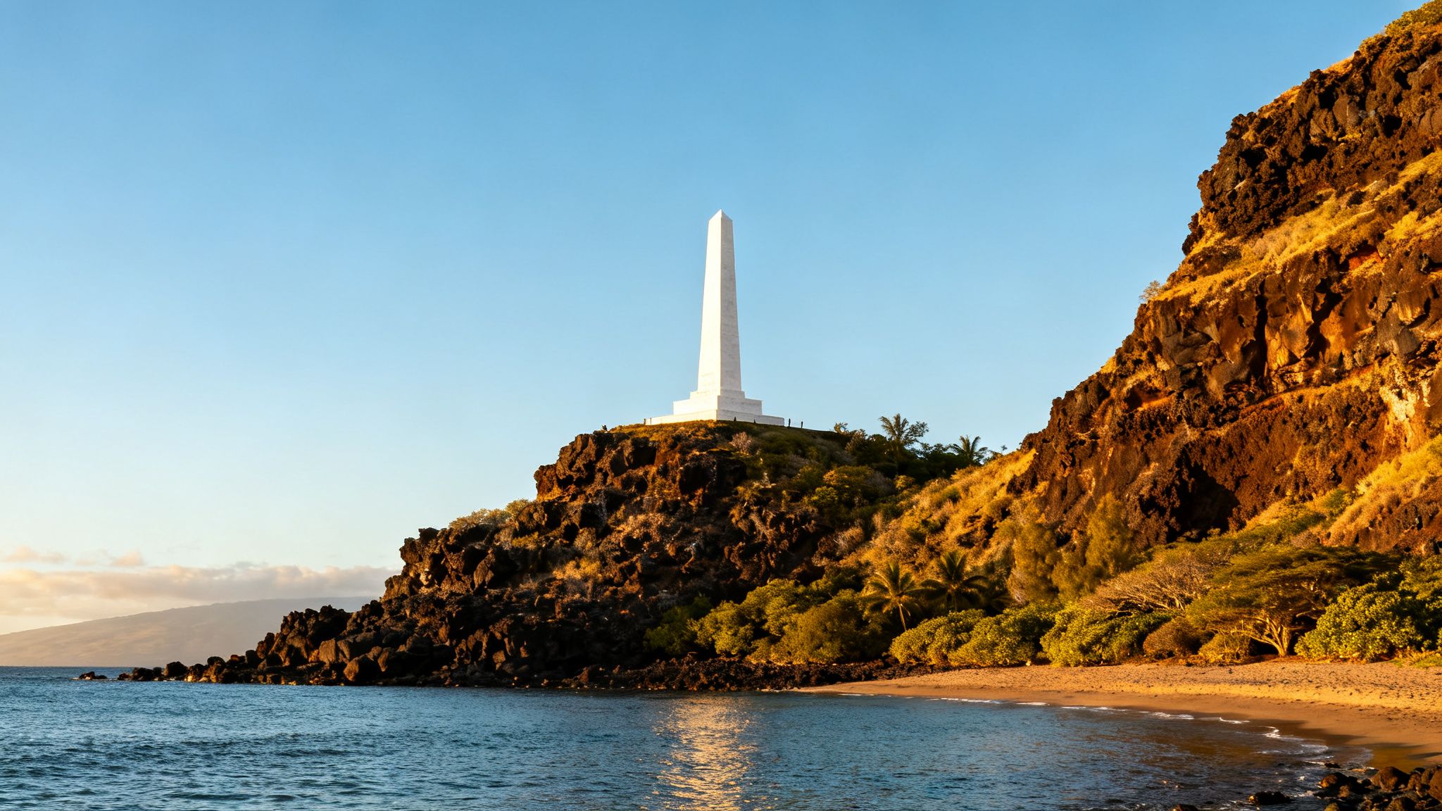 White Captain Cook Monument obelisk on a rocky Hawaiian cliff overlooking a calm bay at sunset.