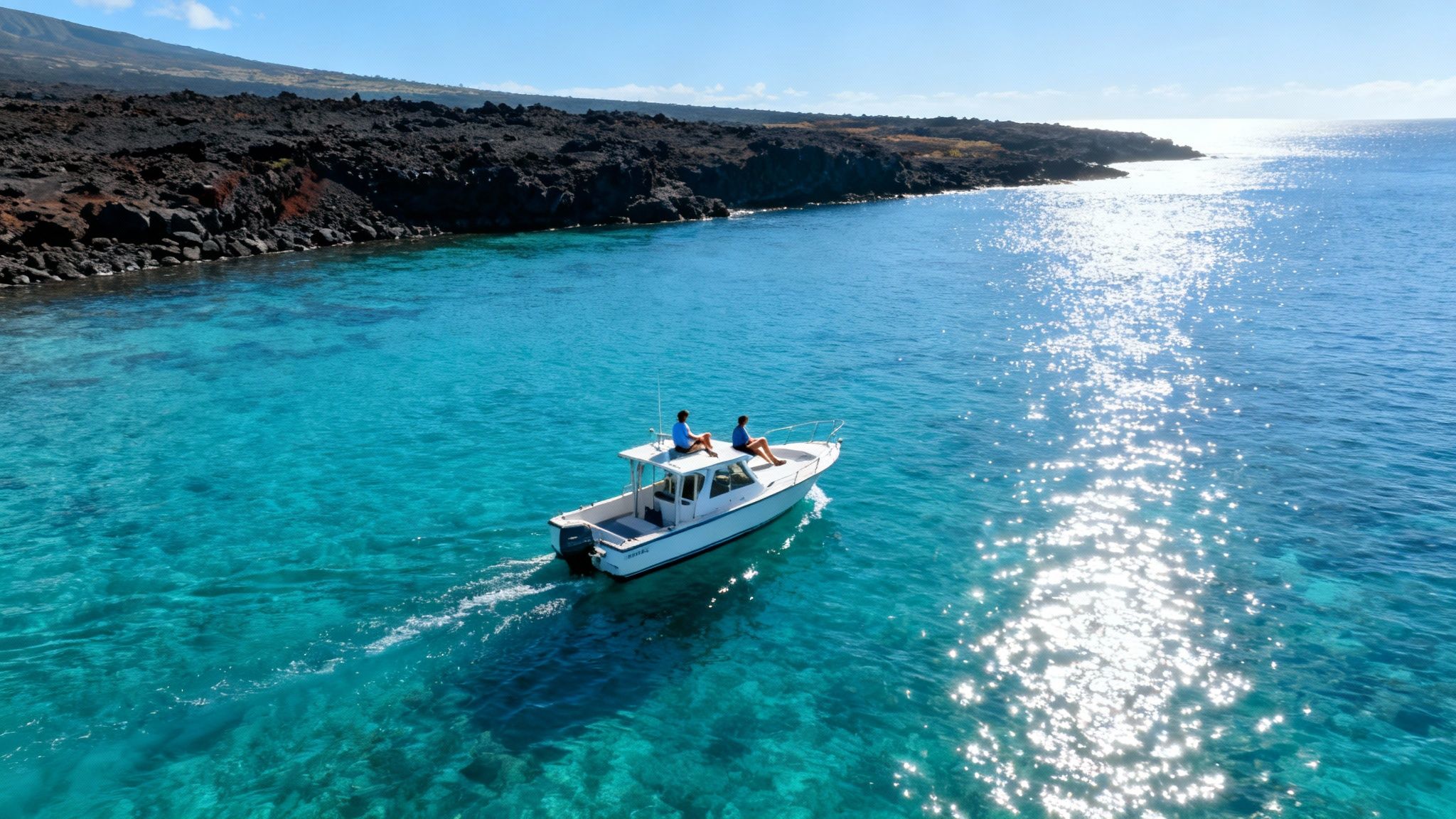 Aerial view of a boat with two people on incredibly clear turquoise water near a dark lava coastline.