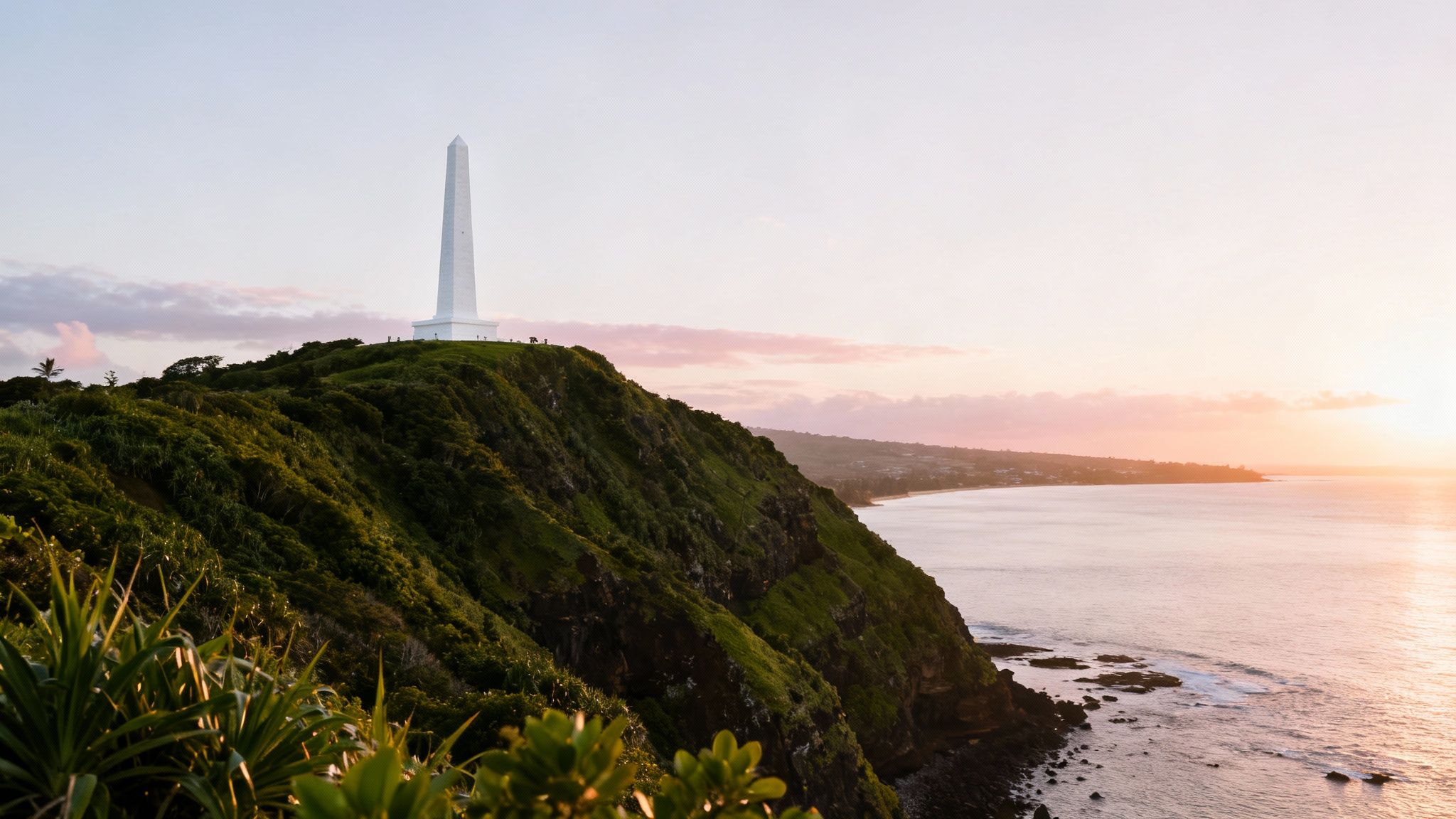 A white obelisk, the Captain Cook Monument, stands on the rocky shoreline of Kealakekua Bay, with lush green cliffs rising behind it and calm, turquoise water in the foreground.