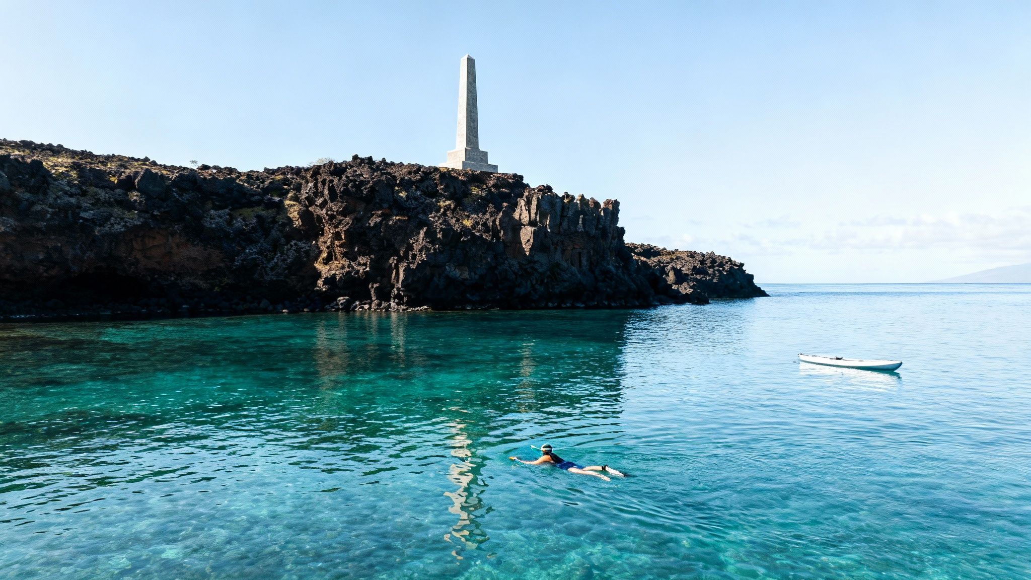 A person snorkeling in crystal-clear turquoise water near a rocky shore with an obelisk monument.