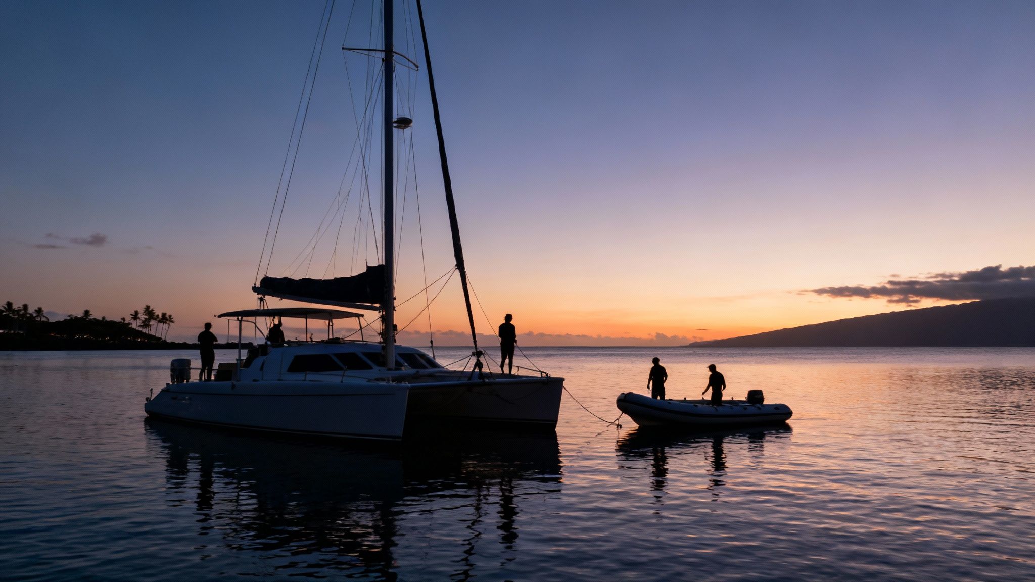 Silhouettes of people on a catamaran and dinghy enjoying a beautiful ocean sunset.