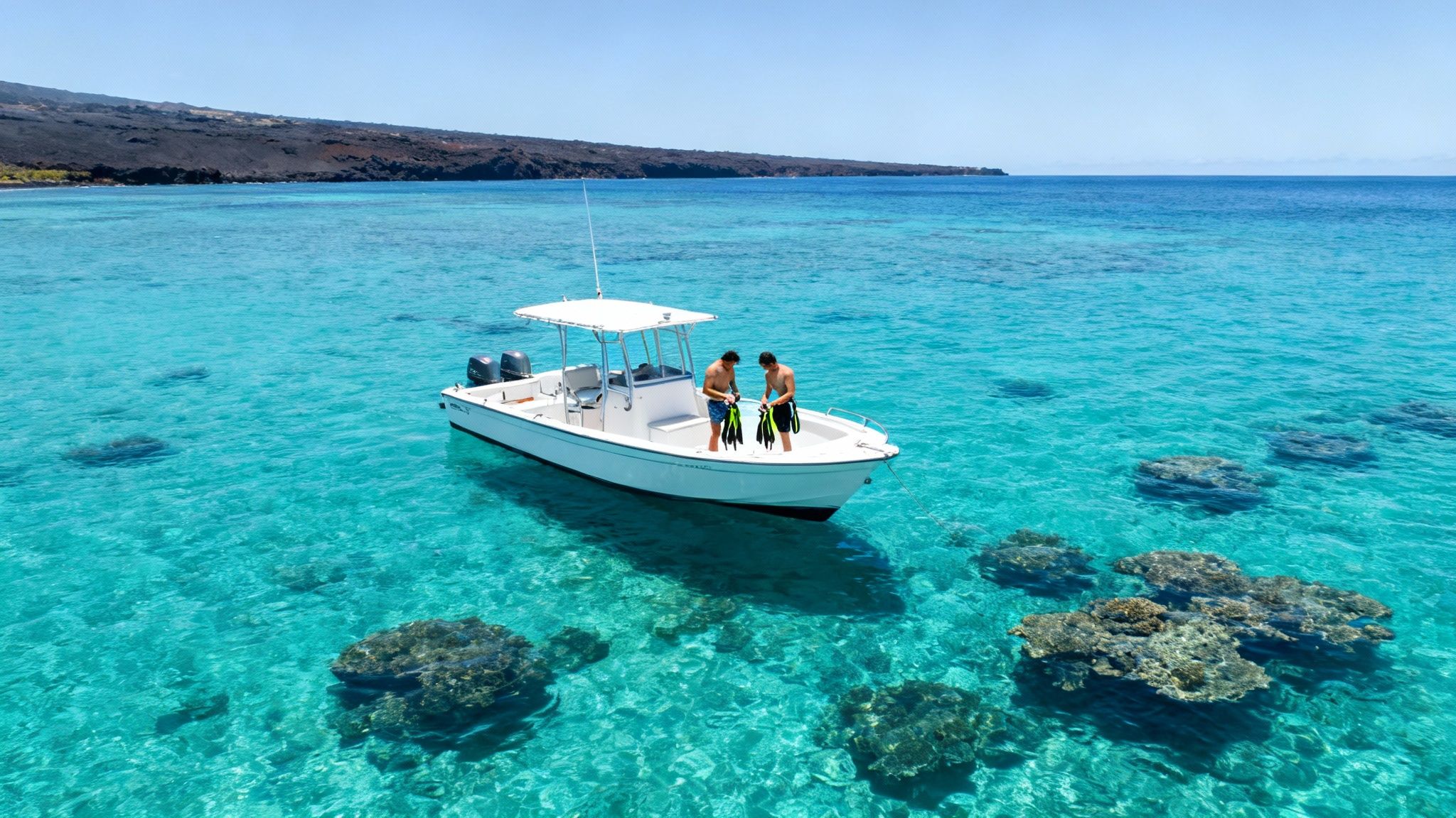 Two men on a white boat in clear turquoise water preparing to snorkel near a volcanic coastline.