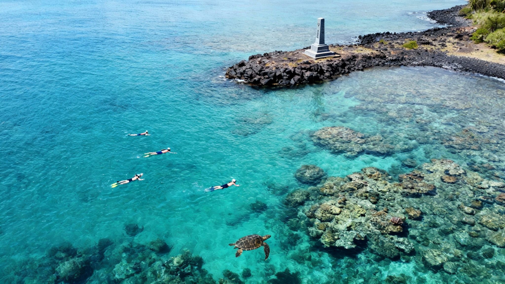 Aerial view of snorkelers, a sea turtle, and coral reefs in clear blue Hawaiian waters near a coastal monument.