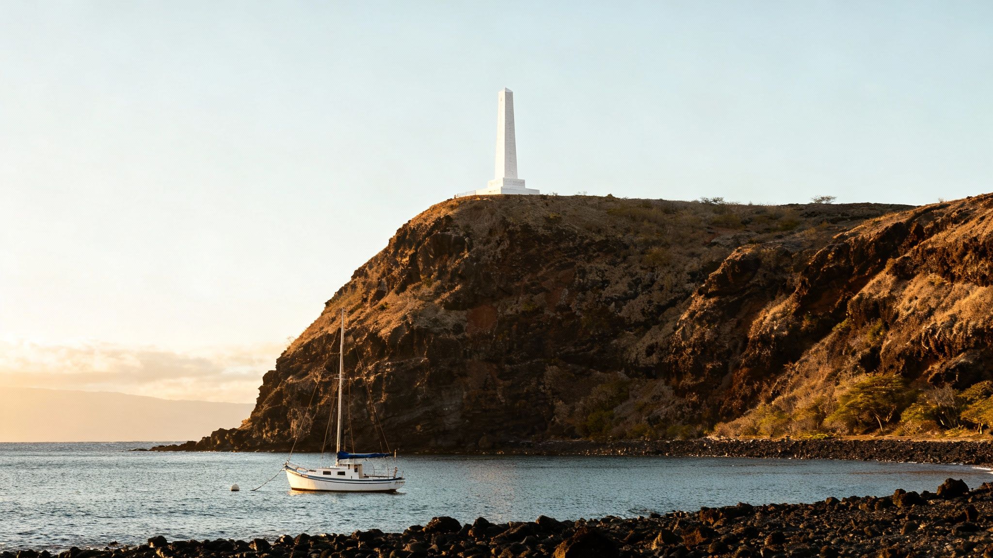 Serene view of a sailboat anchored in a calm bay with a monument atop a rocky cliff.