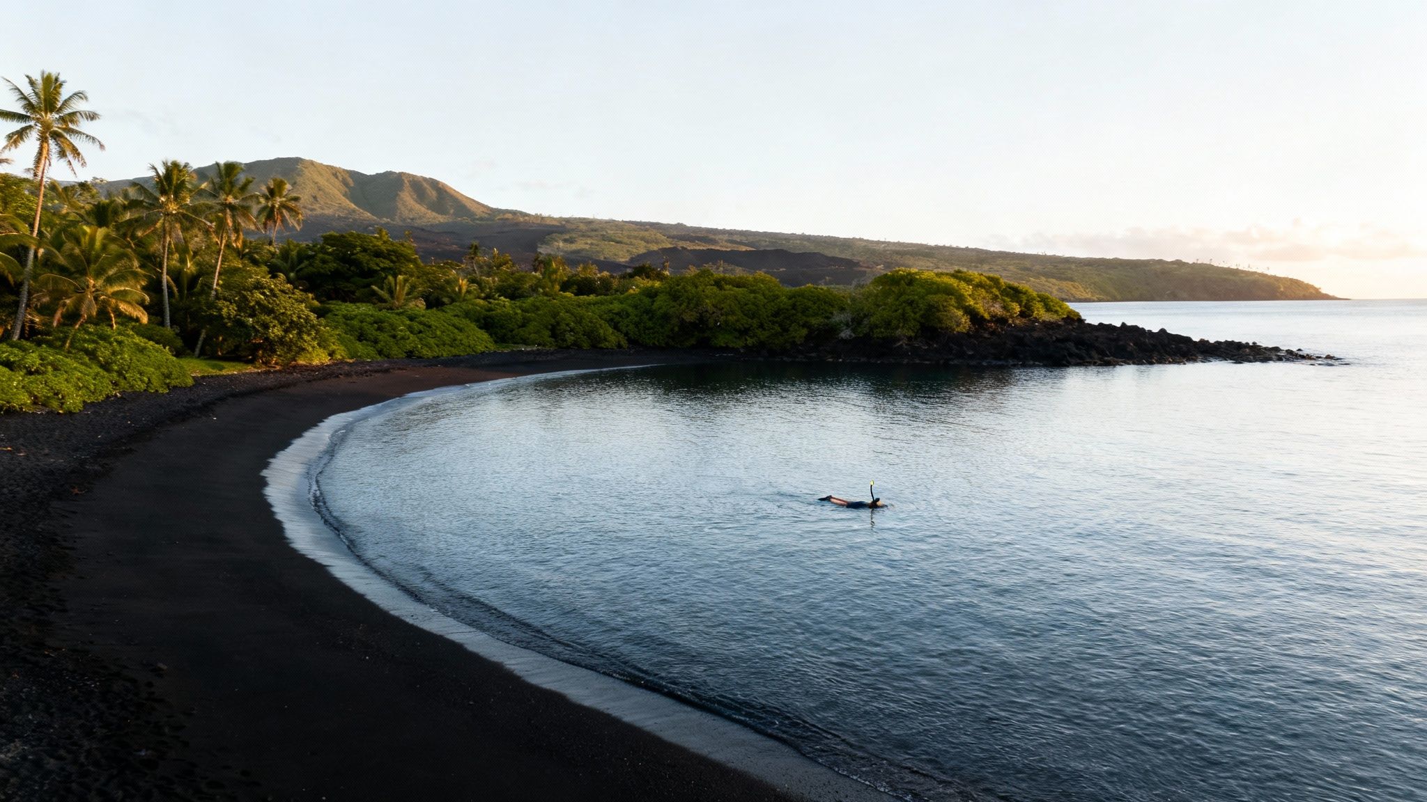 A person snorkels in a calm bay with a black sand beach, lush palm trees, and volcanic mountains at sunrise.
