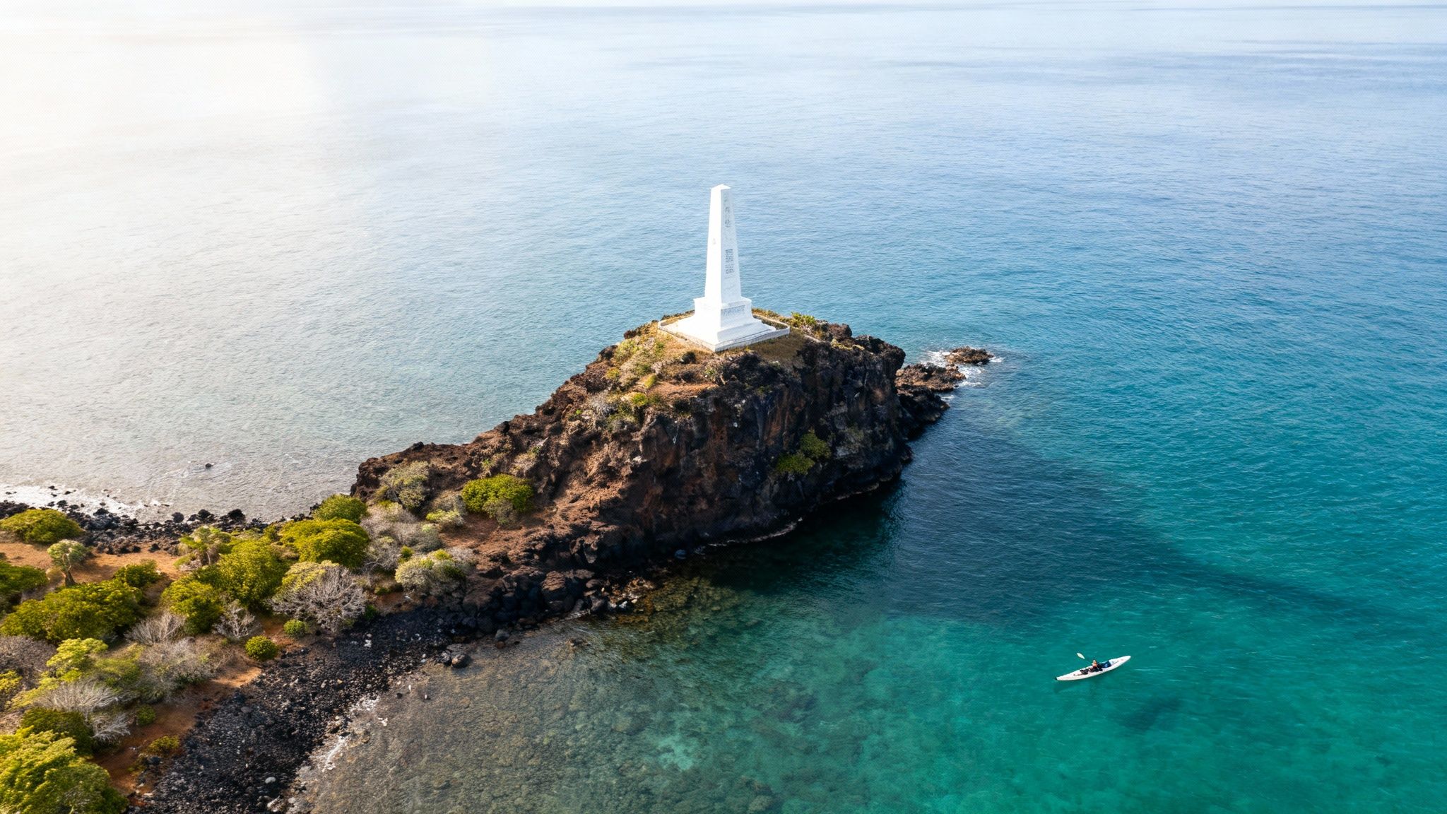 Aerial view of a white obelisk monument on a rocky island surrounded by clear blue ocean, with a kayaker.